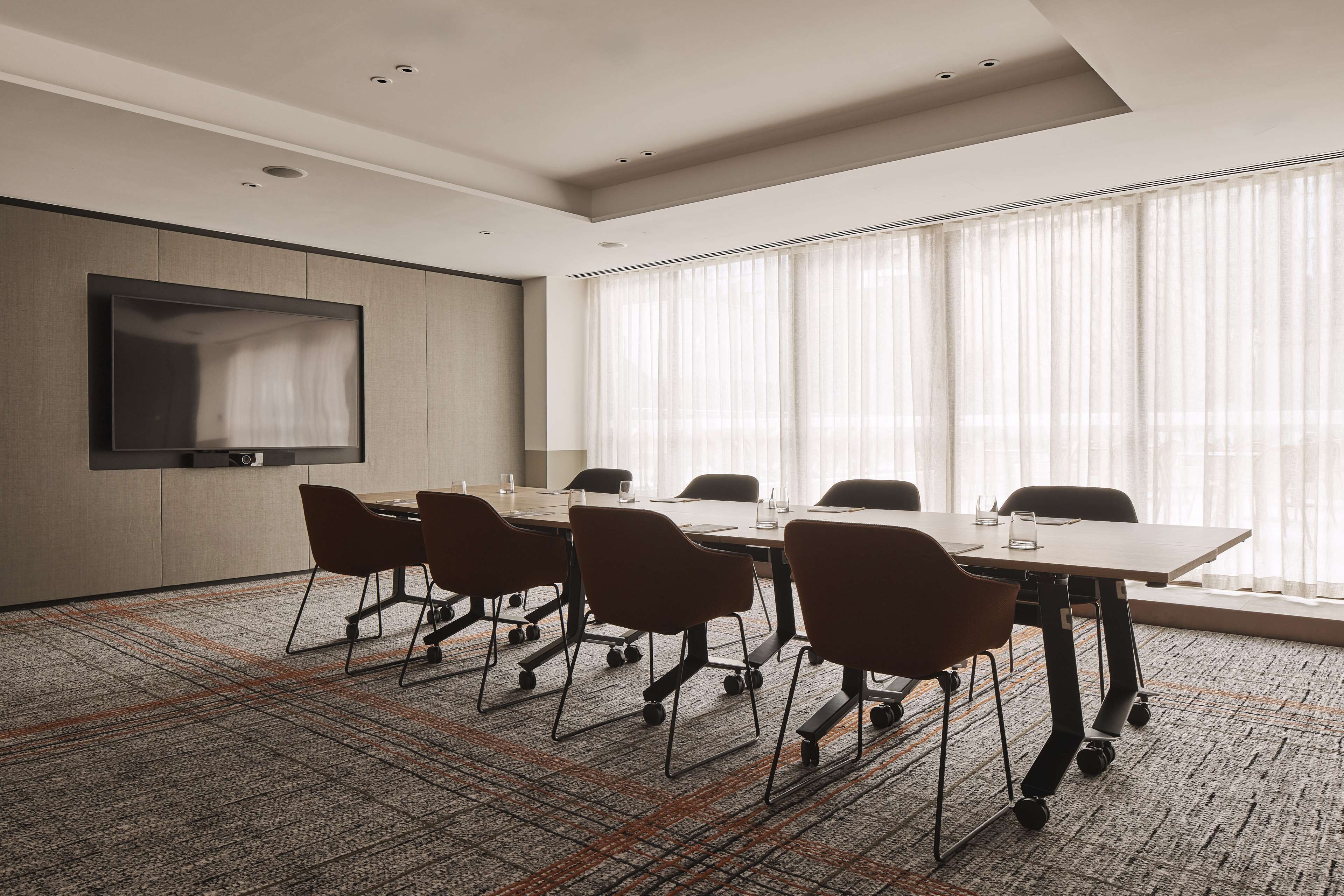 A conference room with a large table, six chairs, a flat-screen TV, and large windows with sheer curtains.
