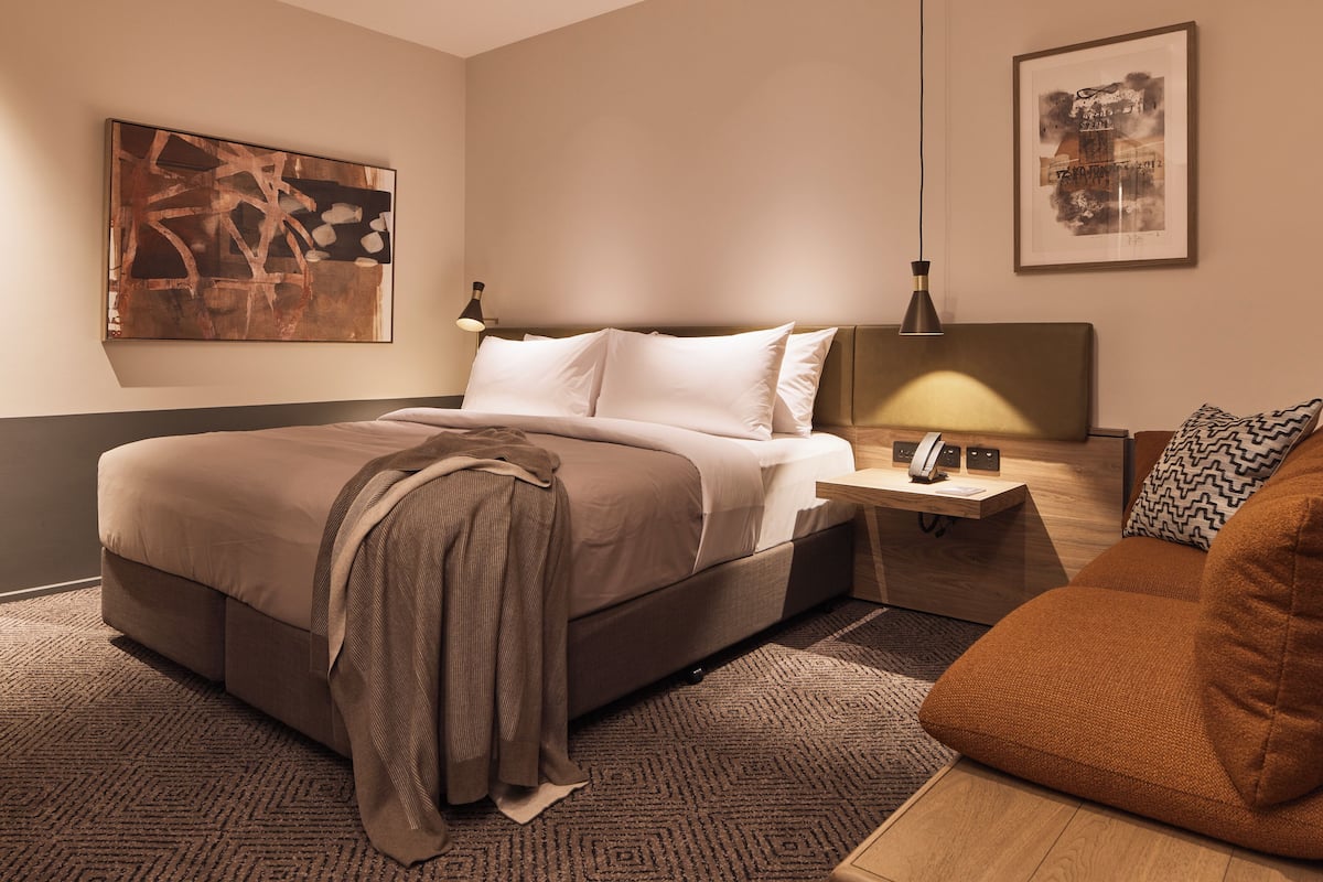 Ein modernes Hotelzimmer mit einem großen Bett, einem Nachttisch mit Lampe, einer kleinen Sitzecke und Bildern an den Wänden.