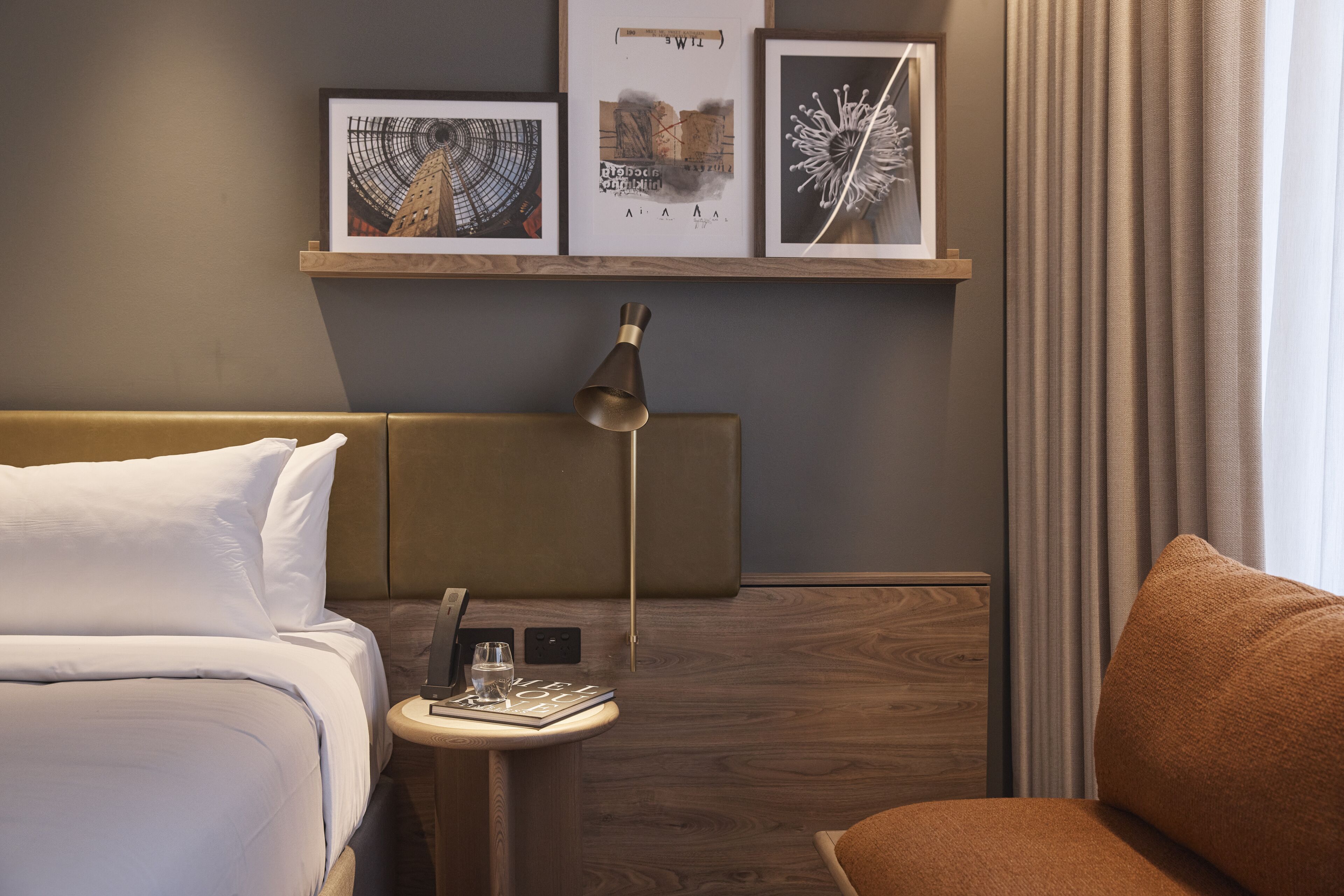 Quarto de hotel com uma cama, uma mesa de cabeceira com abajur e uma pequena mesa com um livro e um copo. Há quadros na parede acima da cama.