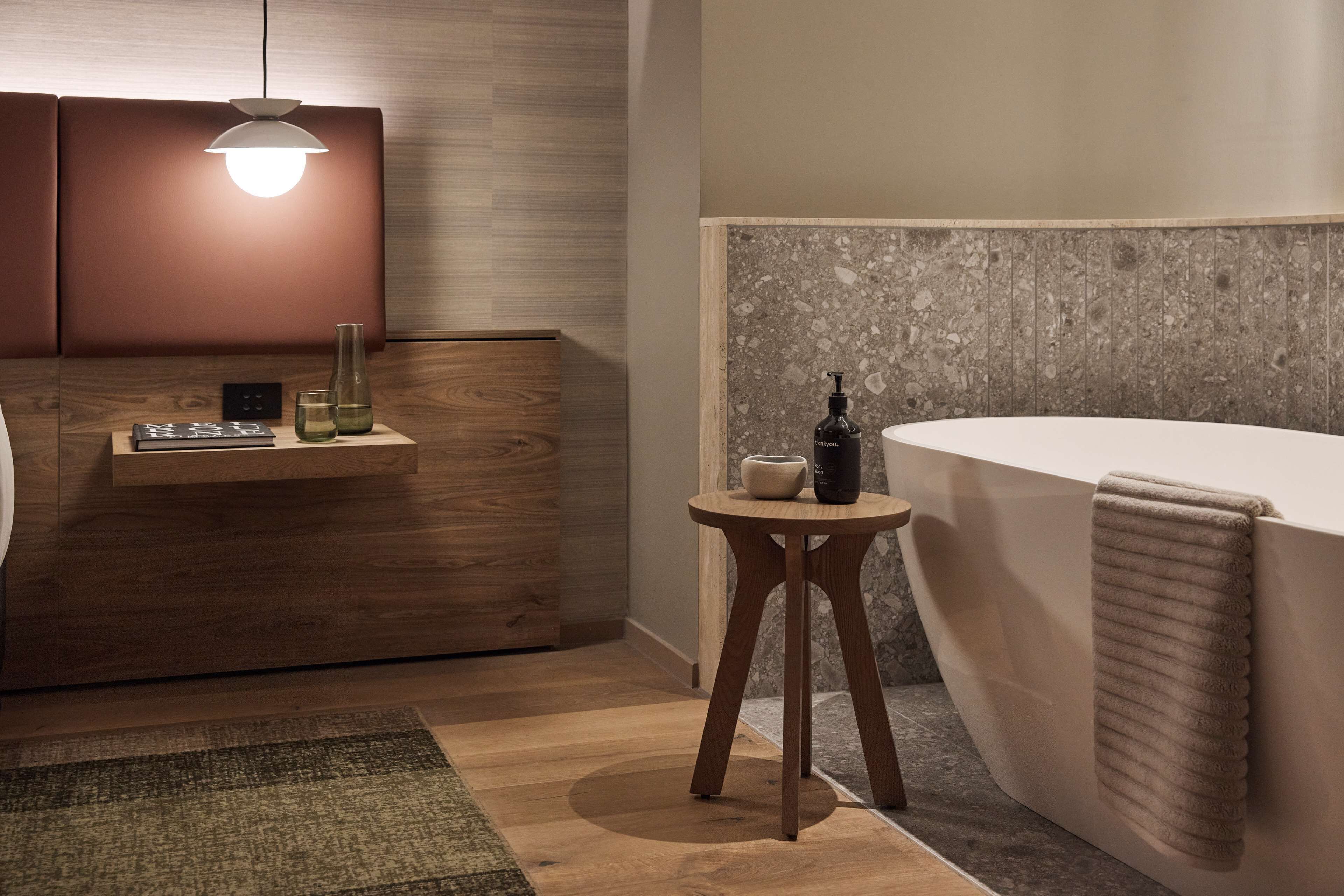 Une salle de bain moderne avec un meuble-lavabo en bois, un tabouret rond en bois, un flacon de savon liquide et une serviette accrochée au mur.