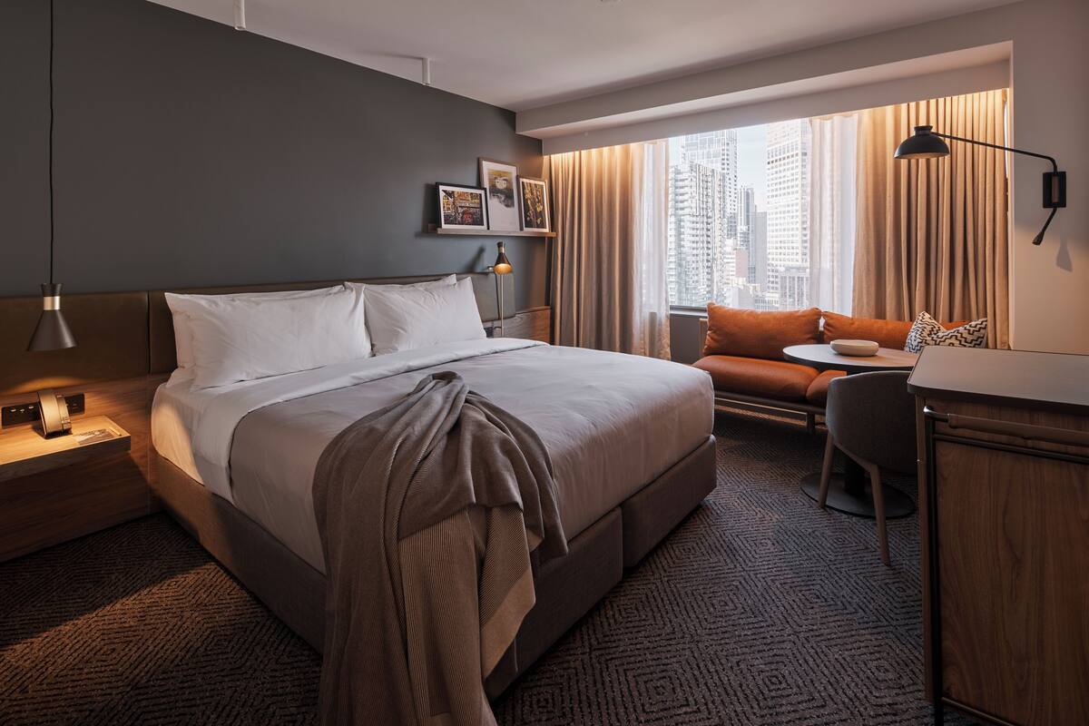 Ein Hotelzimmer mit einem großen Bett, einem Schreibtisch, einem Stuhl und einer Couch. Ein Fenster mit Vorhängen bietet Blick auf die Stadt.