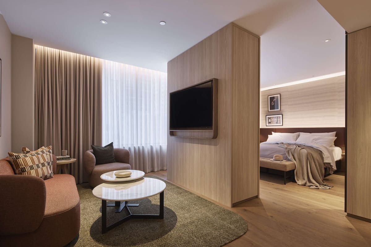 Ein modernes Hotelzimmer mit einer Couch, einem Couchtisch, einem an der Wand befestigten Fernseher, einem Bett mit Kopfteil und einem Nachttisch.