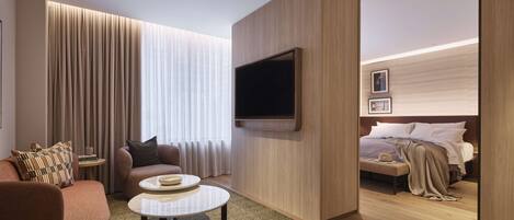 A modern hotel room with a sofa, a coffee table, a TV mounted on the wall, a bed with a headboard, and a bedside table.