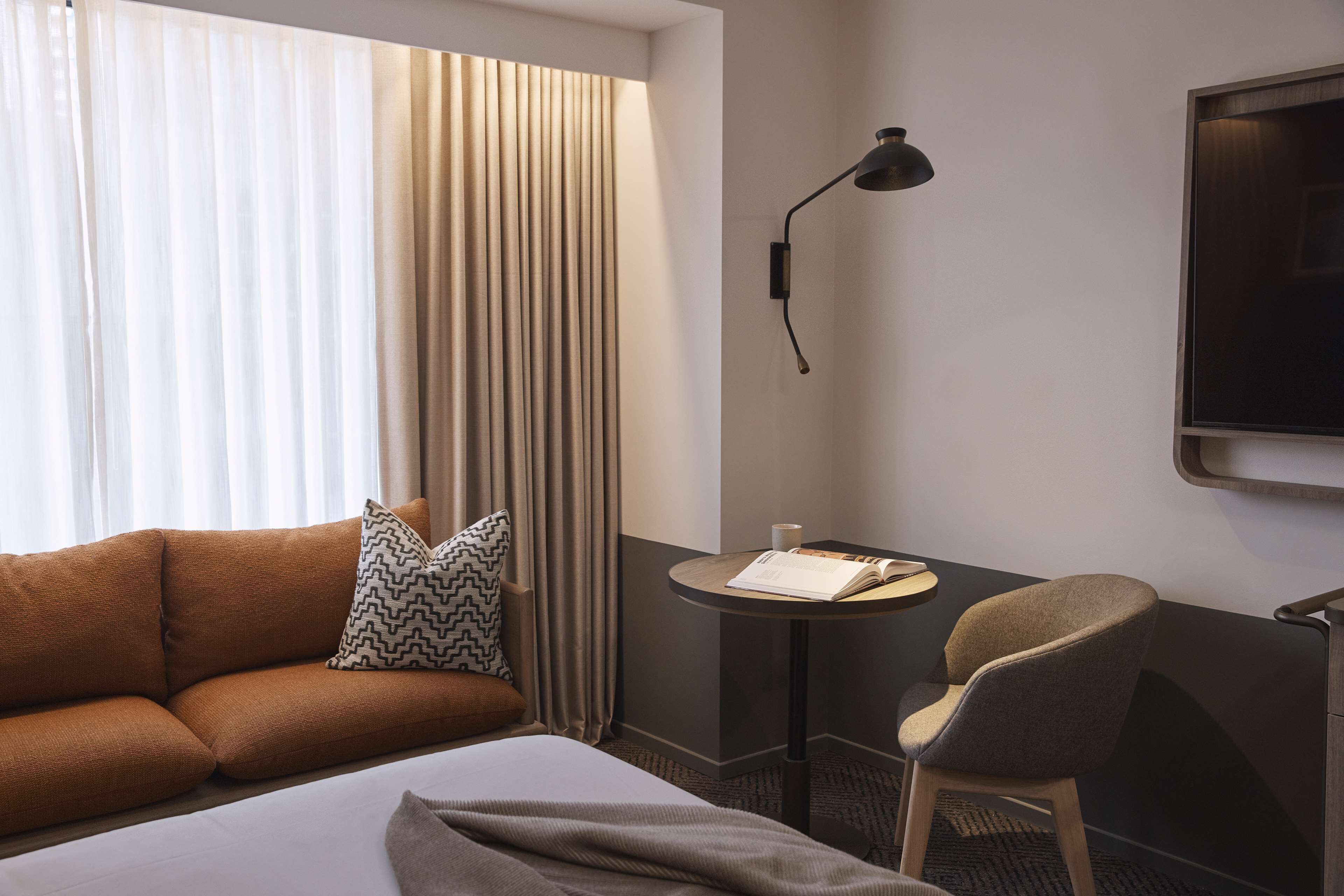 A hotel room with a bed, a sofa, a chair, a small table with a book and a cup, a lamp, and a TV on the wall.