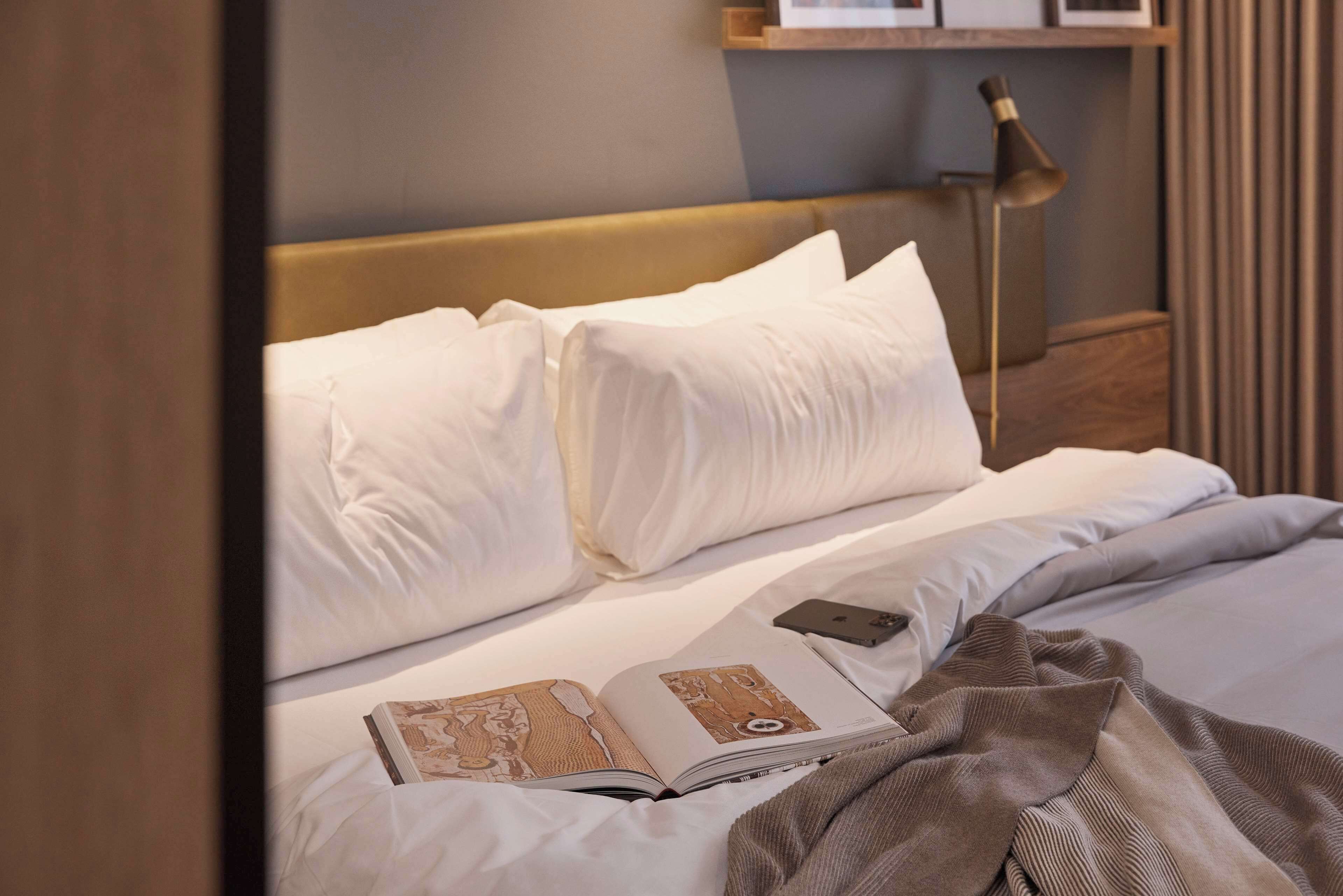 A neatly made bed with white linens, a book, and a smartphone on it.