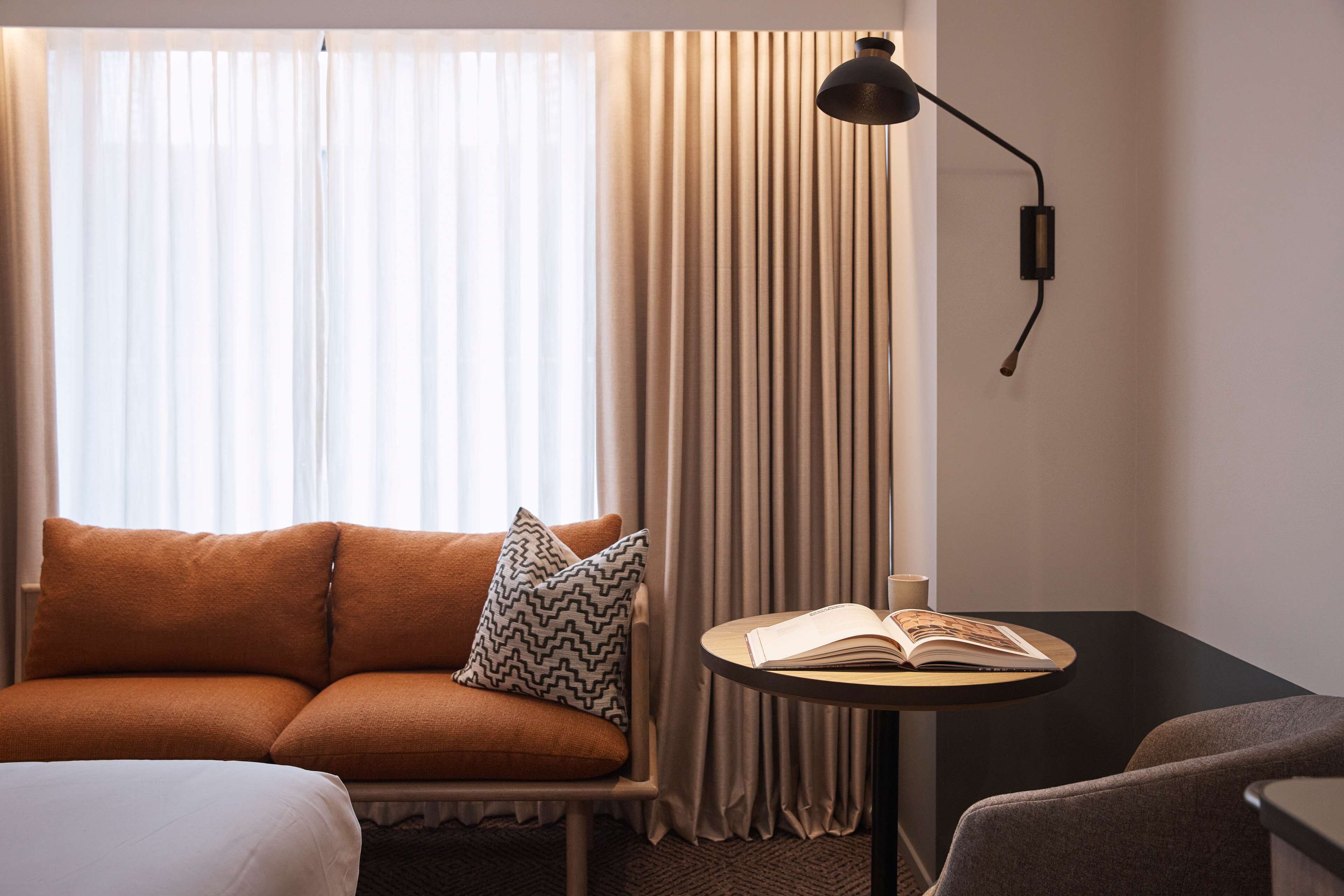 A hotel room with a sofa, a small table with an open book, a lamp, and curtains.