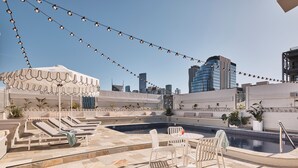 A rooftop pool area with lounge chairs and a covered seating area, surrounded by city buildings.
