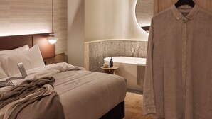 A modern hotel room with a bed, a laptop, a bedside table with a bottle, and a hanging shirt.