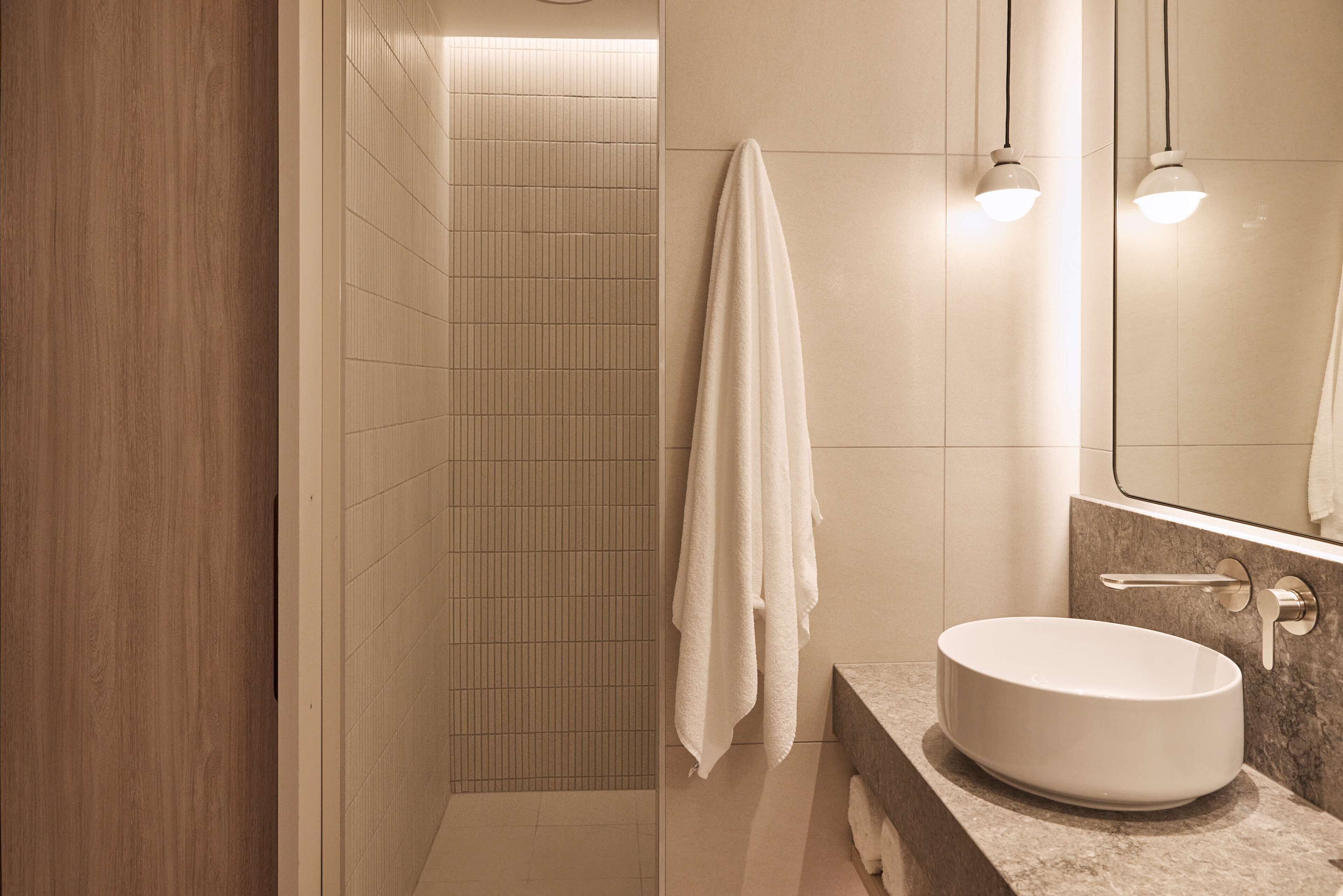 A modern bathroom with a wooden wall, a tiled shower area, a white sink, and a towel hanging on the wall.