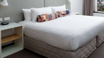 Una cama bien hecha con una colcha acolchada, una almohada blanca y una almohada colorida. Hay una mesita de noche con una lámpara y un enchufe. La habitación tiene vista a la ciudad a través de una ventana con cortinas.