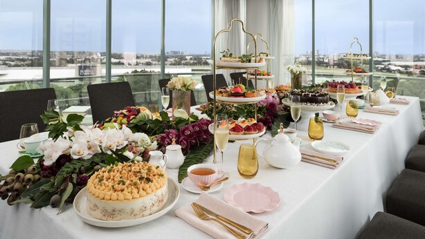 Habitación de hotel en un rascacielos con vistas panorámicas de la ciudad, una amplia mesa de comedor dispuesta con diversos postres y un centro de mesa floral.