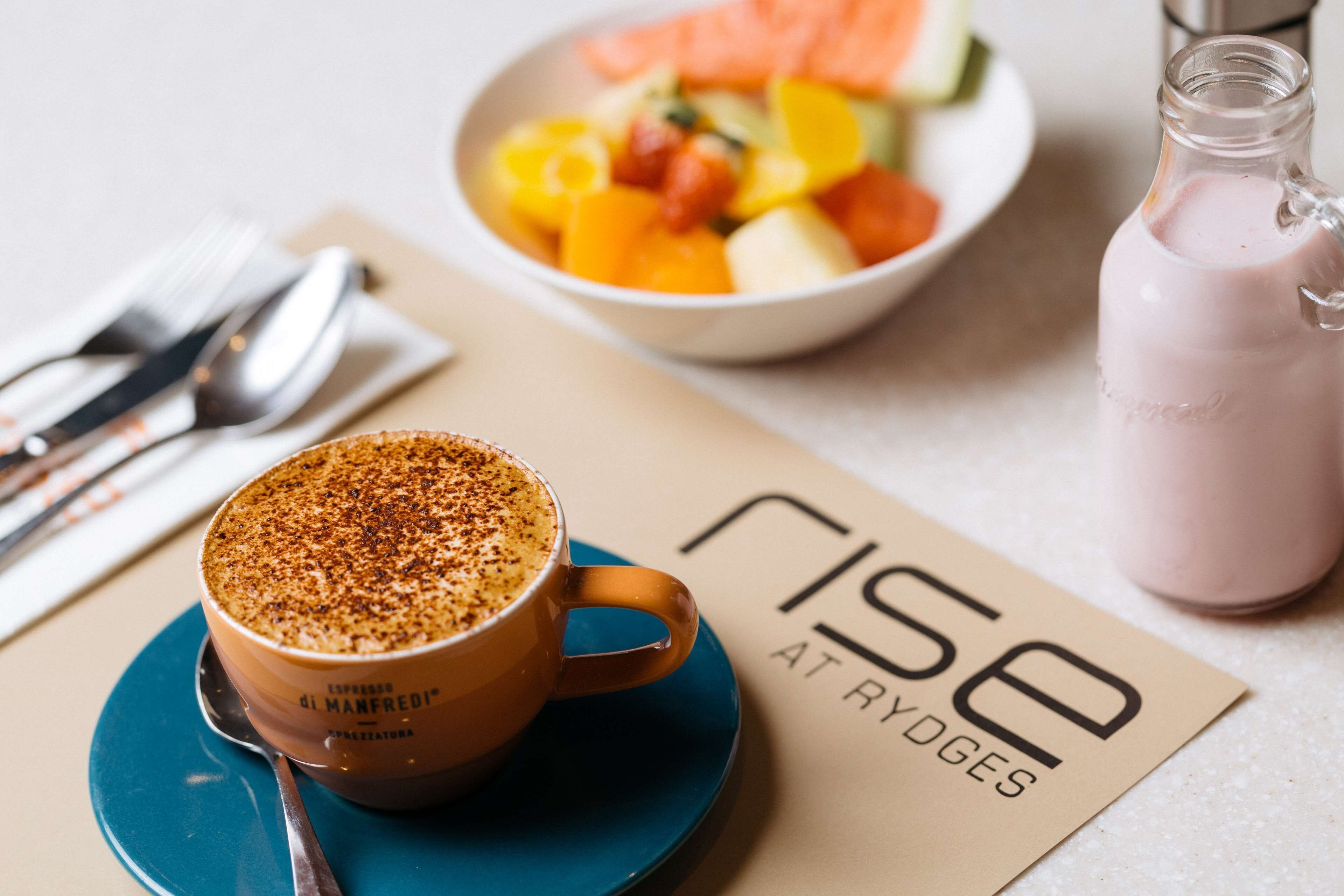 A coffee cup on a blue saucer, a bowl of fruit salad, and a bottle of smoothie on a table with a napkin and cutlery.