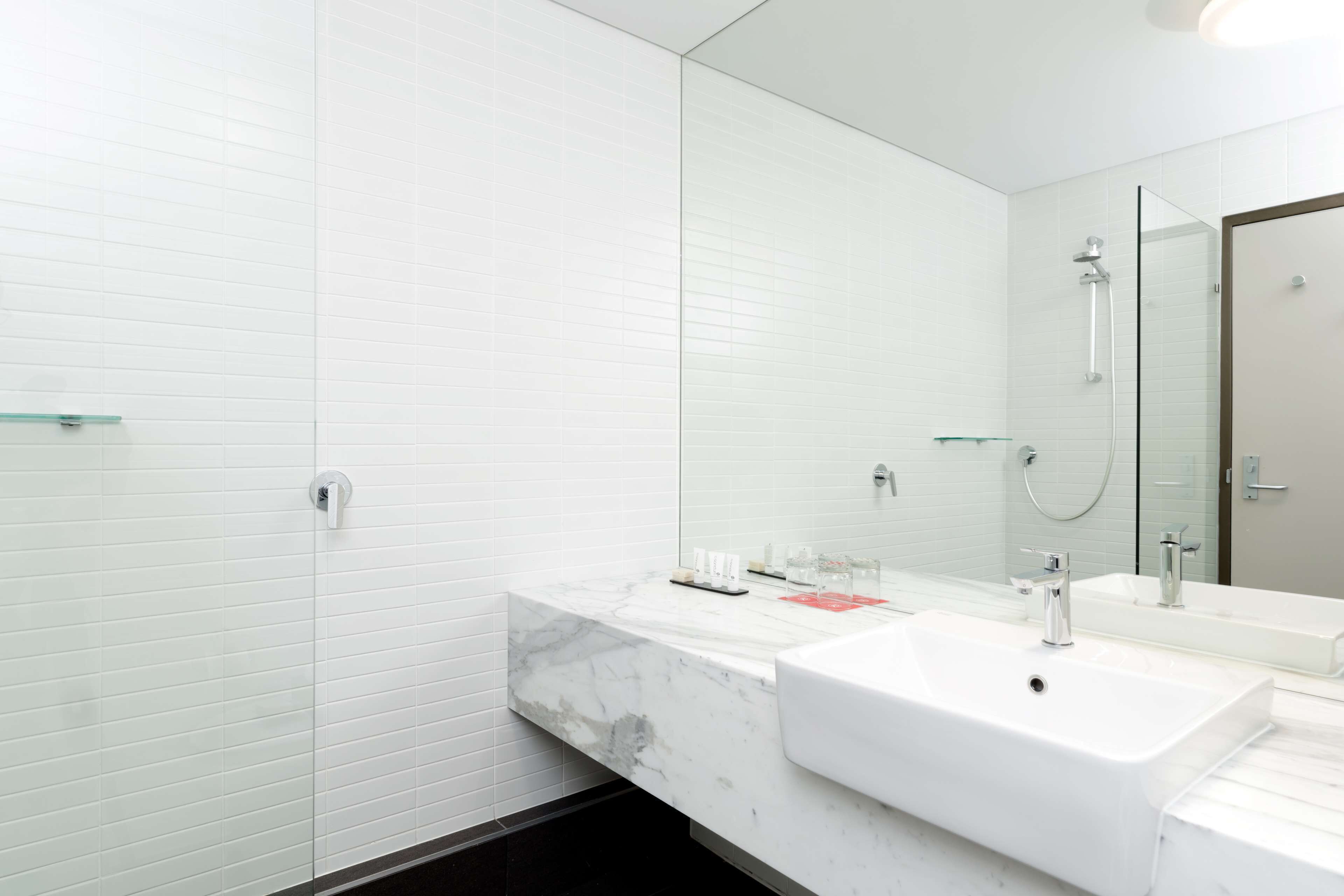 Une salle de bain moderne avec des murs recouverts de carreaux blancs, un grand miroir, un lavabo blanc et une douche en verre.