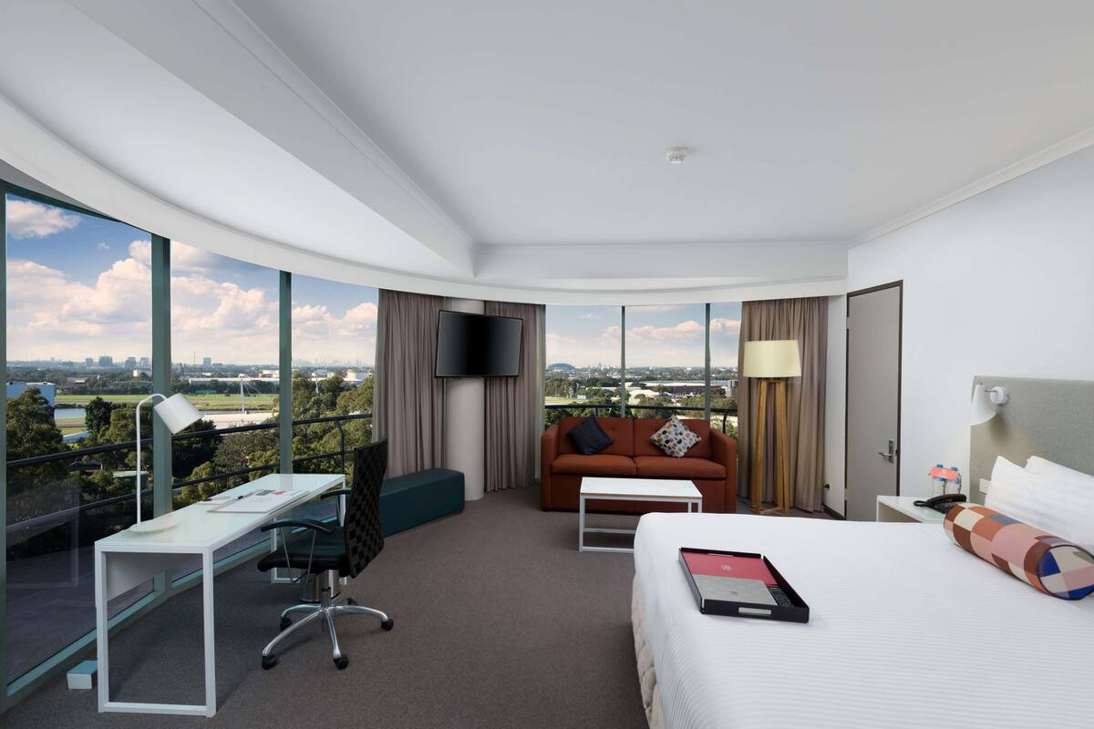 Una habitación de hotel moderna con escritorio, un sofá cómodo, una cama y un amplio ventanal con vistas a la ciudad.