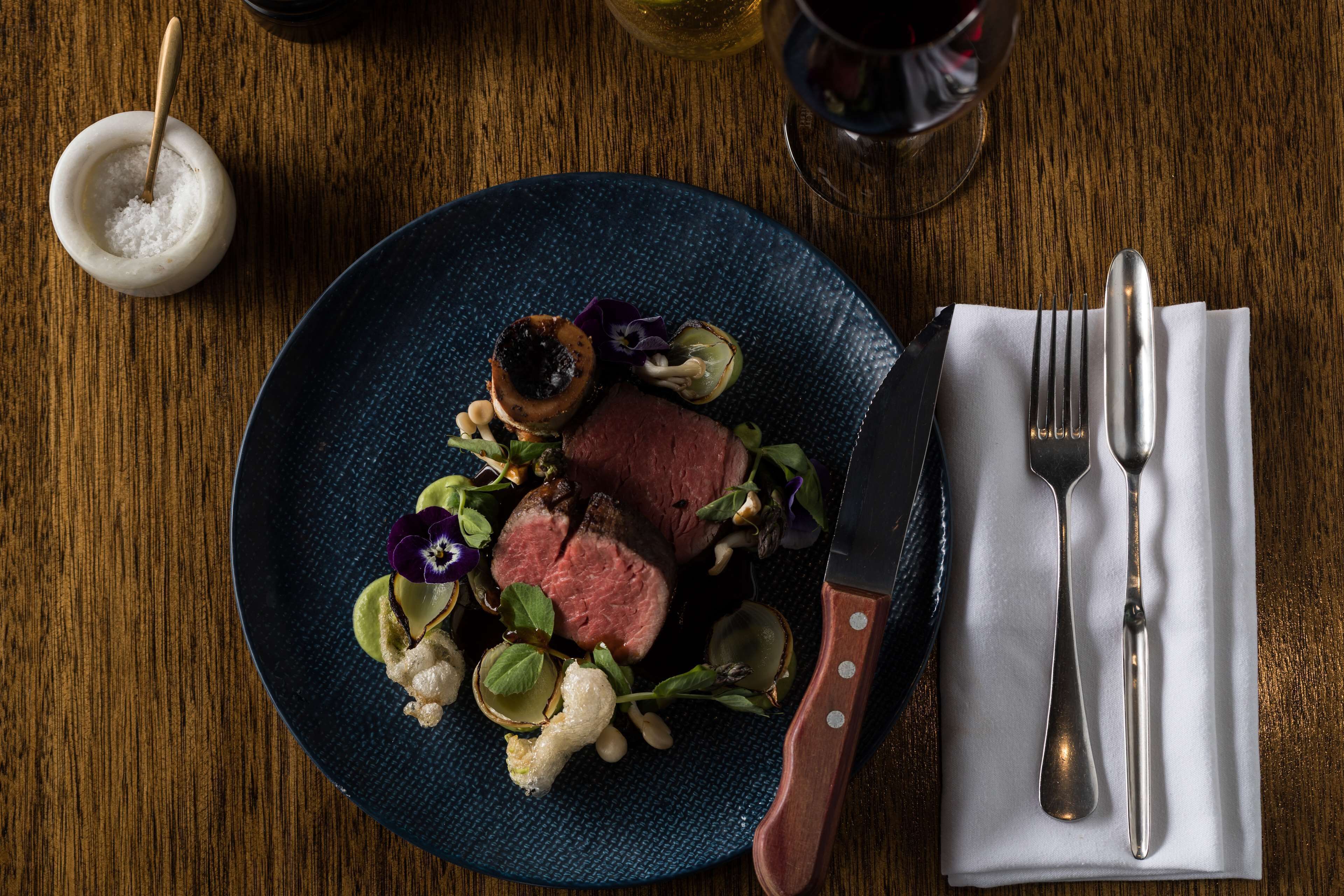 A plate of food with a knife and fork on a napkin, a salt shaker, and a glass of wine on a wooden table.