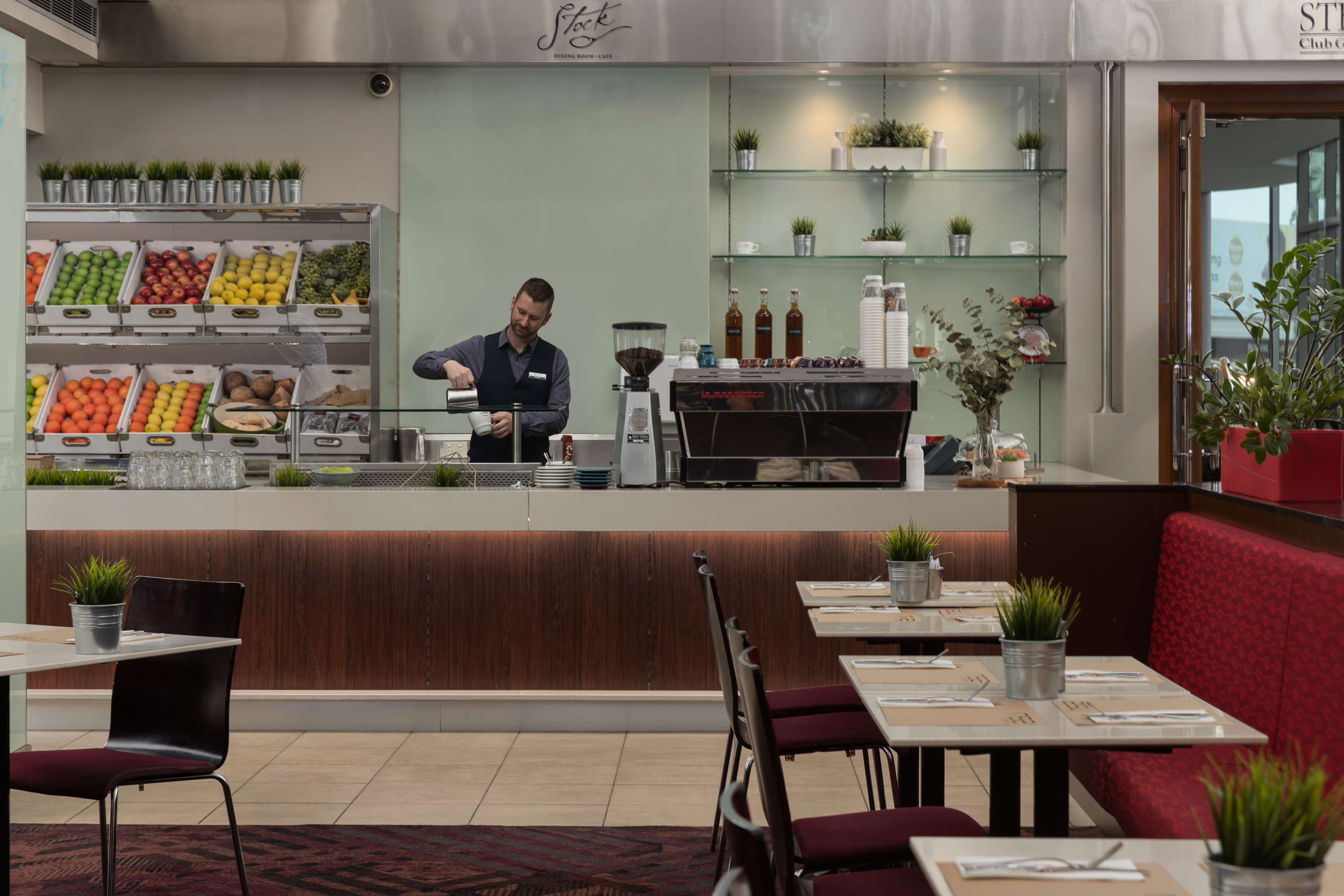 A modern cafe with a barista preparing drinks, a variety of fresh fruits and vegetables on display, and seating areas with potted plants.