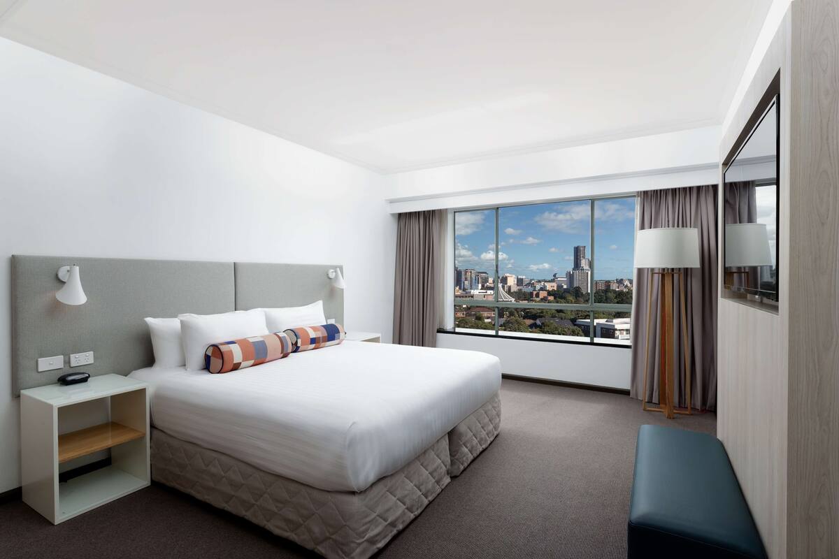Una habitación de hotel moderna con una cama grande, una mesita de noche, un banco y vistas a un paisaje urbano a través de un ventanal amplio.