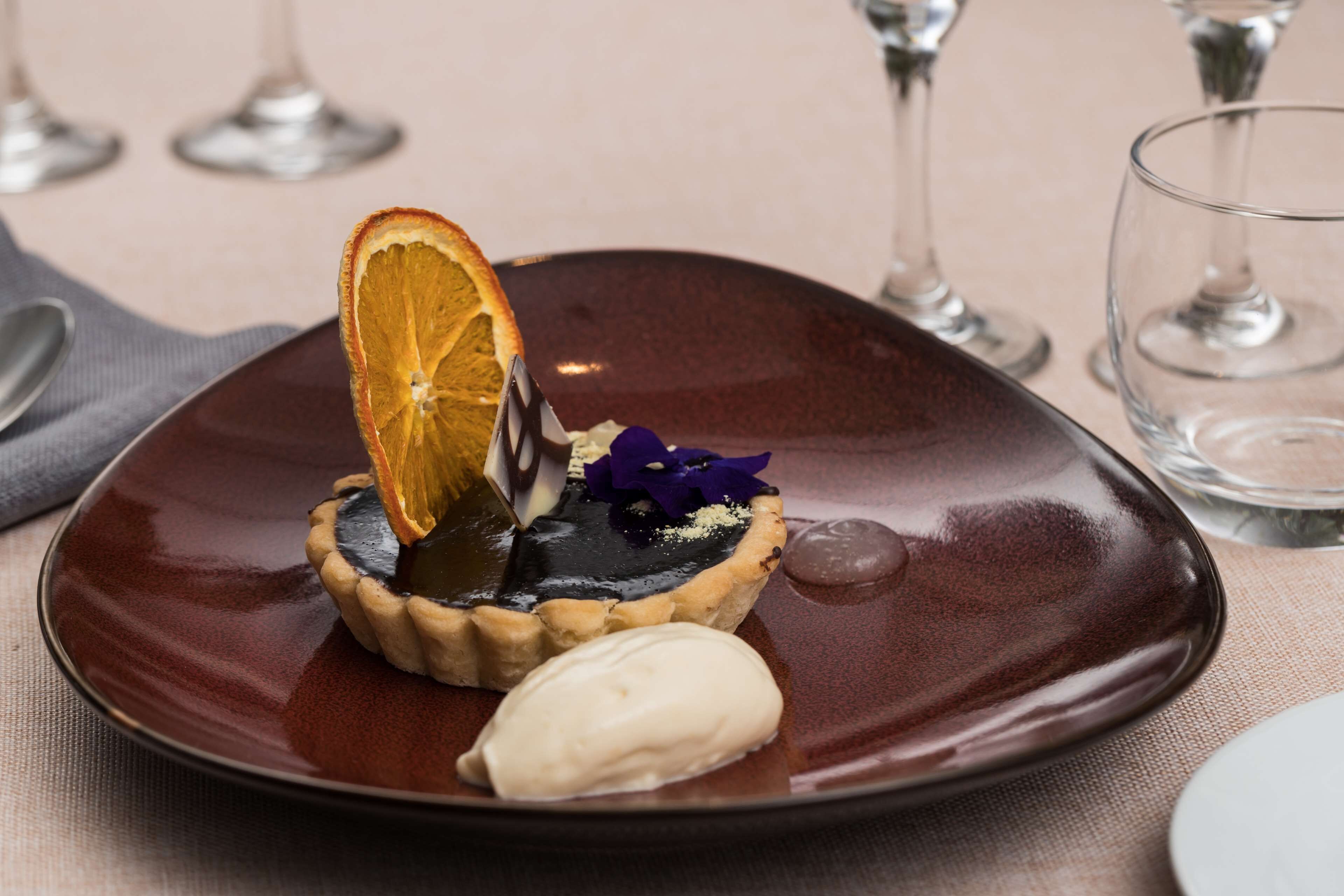 A dessert on a plate with a dark brown rim, a slice of orange, and a small purple flower.