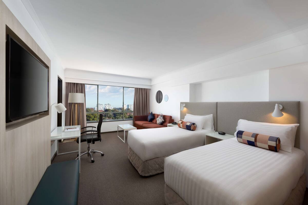Habitación de hotel con dos camas, un escritorio, televisión y un ventanal con vistas a la ciudad.
