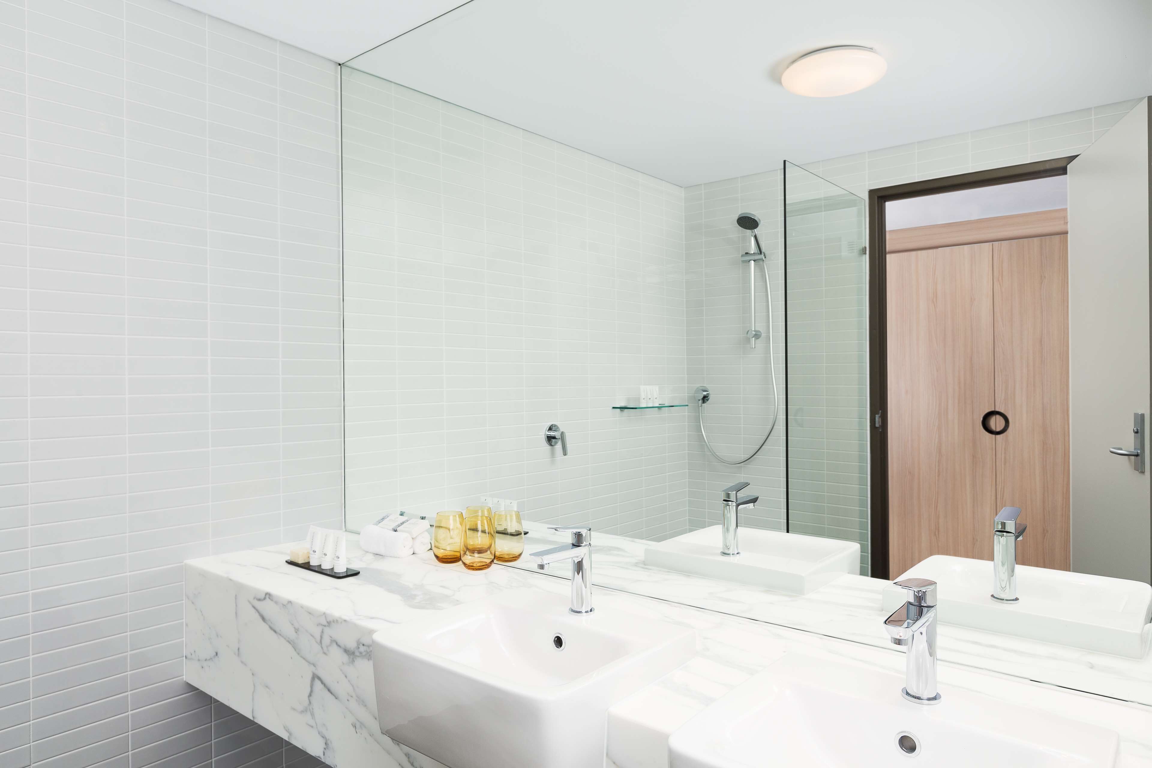 A modern bathroom with a large mirror, two sinks, a shower, and a wooden door.