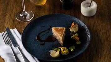 Un plato de postre con una porción de pastel, salsa de chocolate y adornos sobre un plato oscuro.