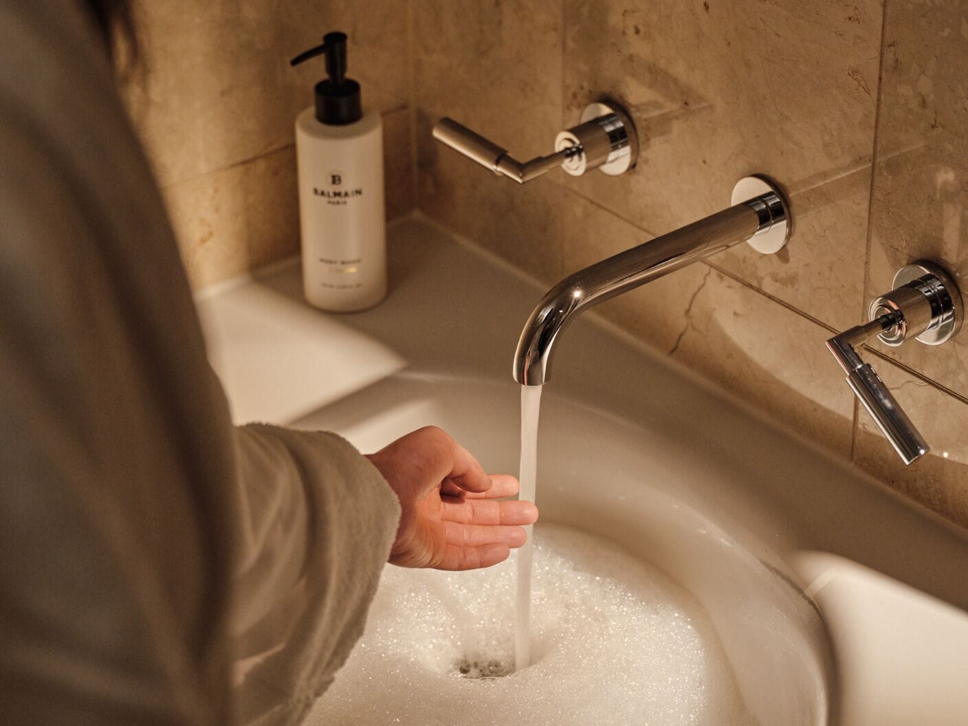 A hand under a running faucet in a bathroom with a bottle of hand soap on the wall.