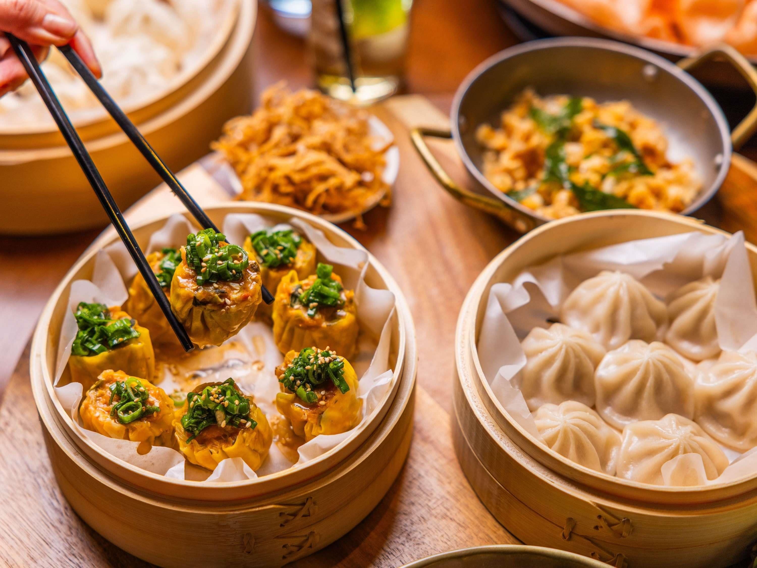 A variety of Asian dumplings in bamboo steamers, a bowl of noodles, and a pan with stir-fried vegetables.