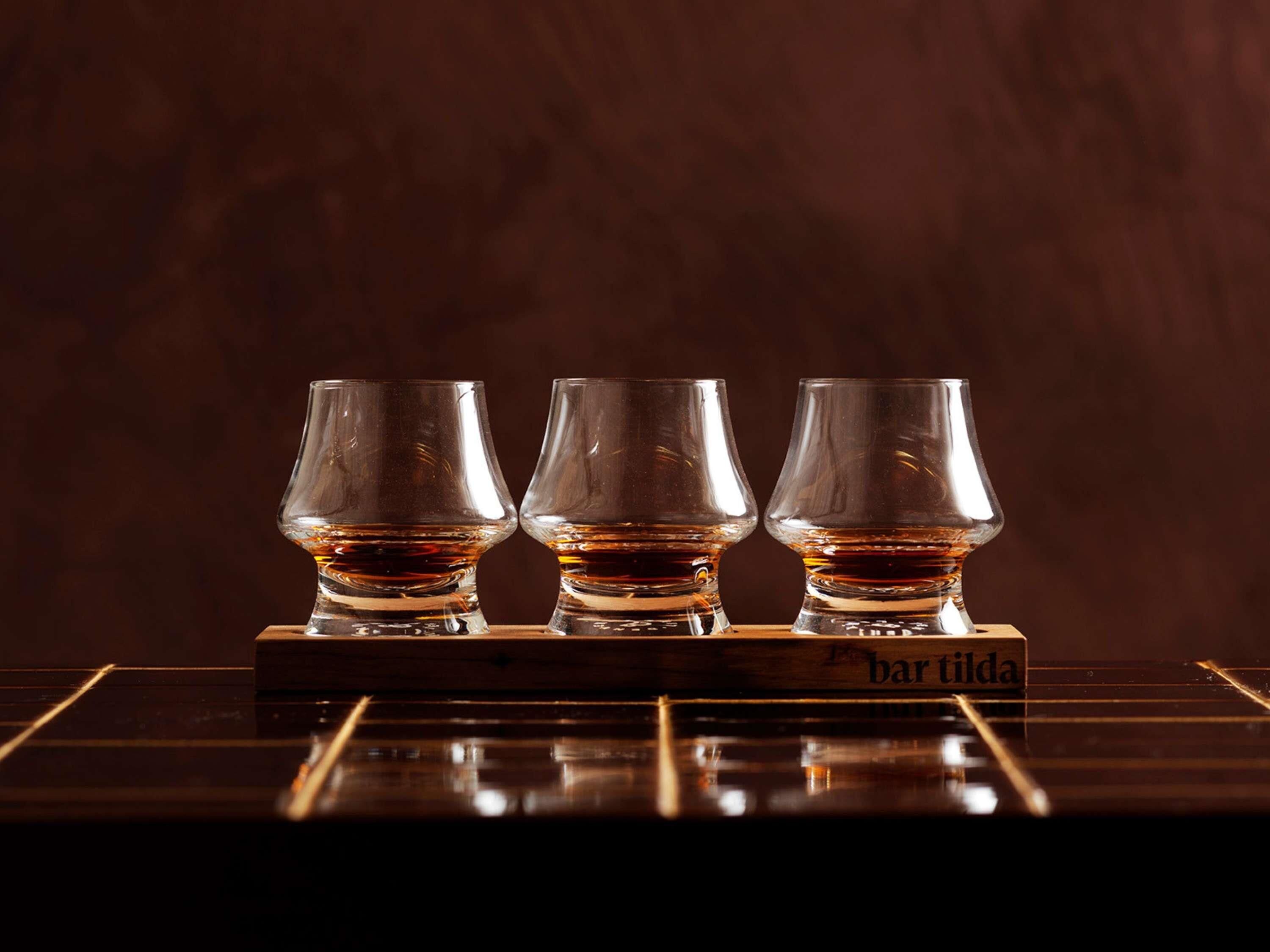 Tres vasos de whisky sobre un tapete de madera con la leyenda 'bar tilda' en una superficie reflectante.