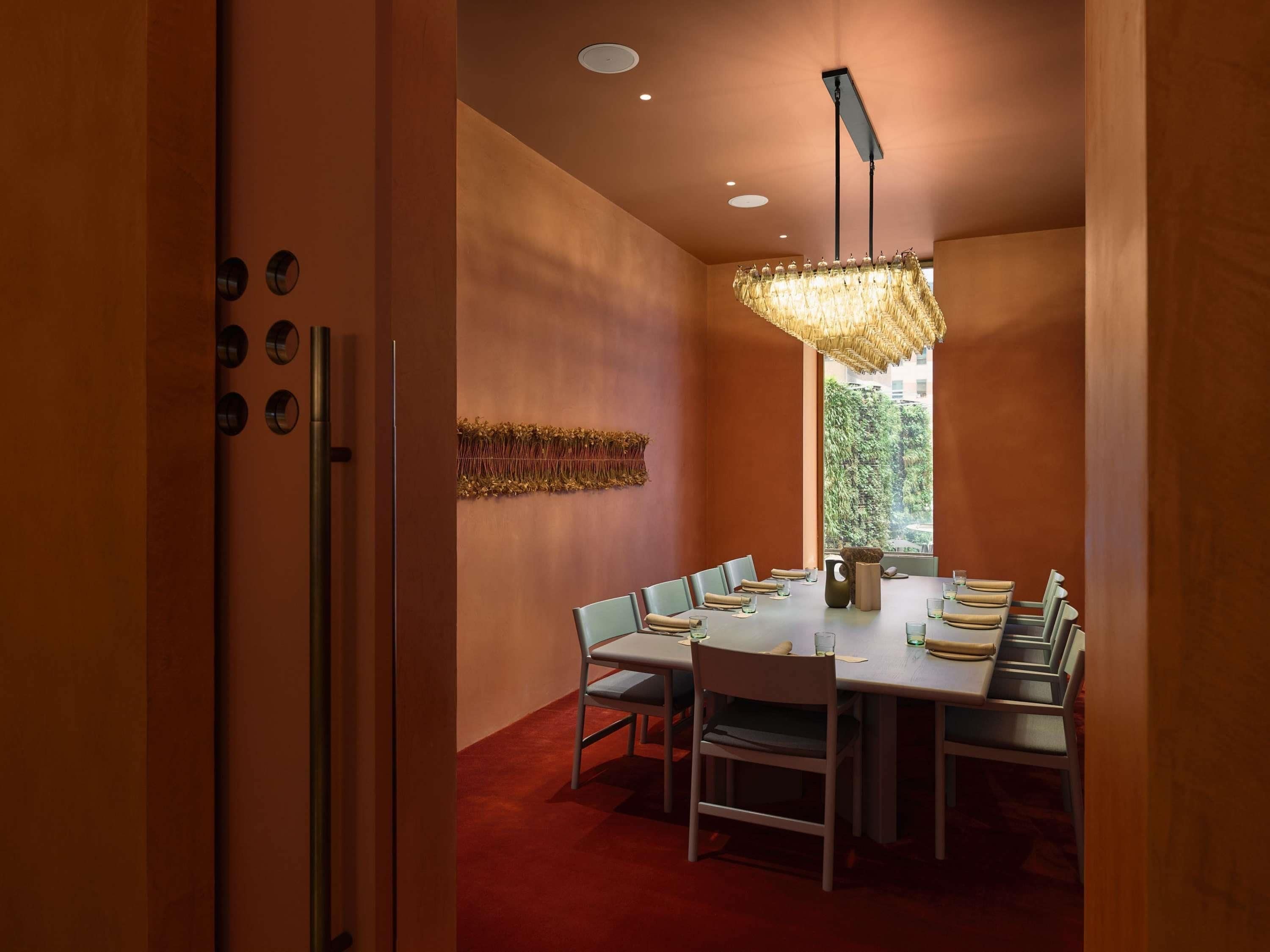 A dining area with a long table, chairs, a chandelier, and a view through a window.