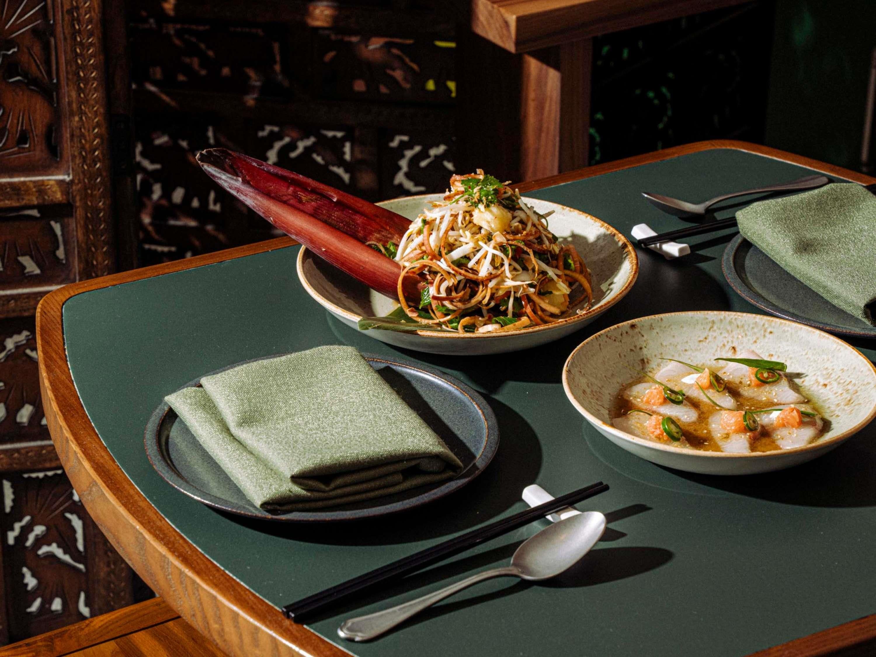 A dining table with three bowls of food, chopsticks, a spoon, and a folded napkin.