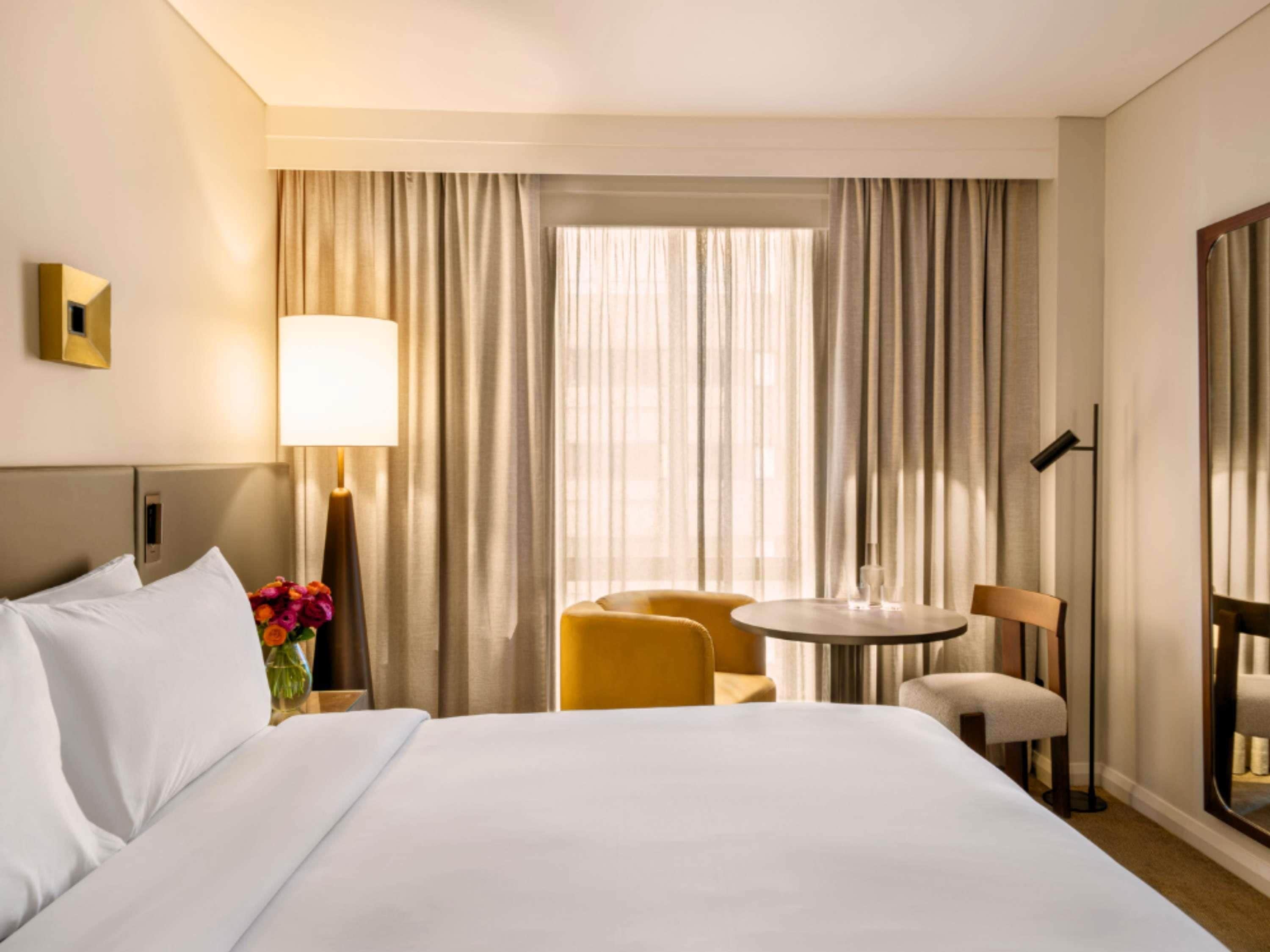 A hotel room with a bed, a desk, a chair, a lamp, and a window with curtains.