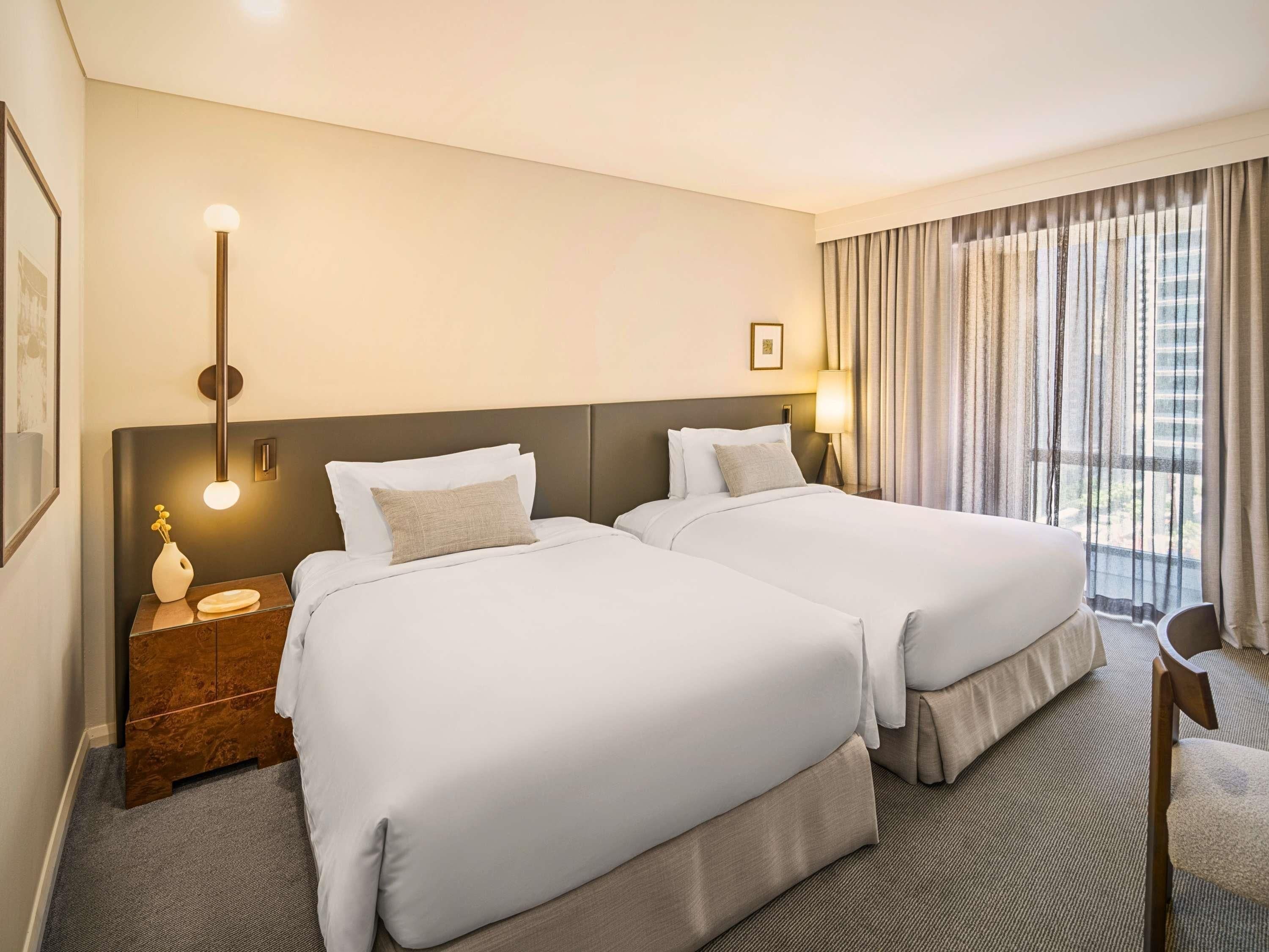 A hotel room with two beds, a nightstand, a lamp, a chair, and a window with curtains.