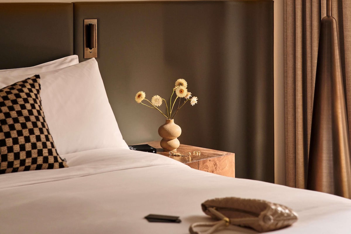 A neatly made bed with white linens, a checkered pillow, a vase with flowers, a small wooden nightstand, and a smartphone on the bed.