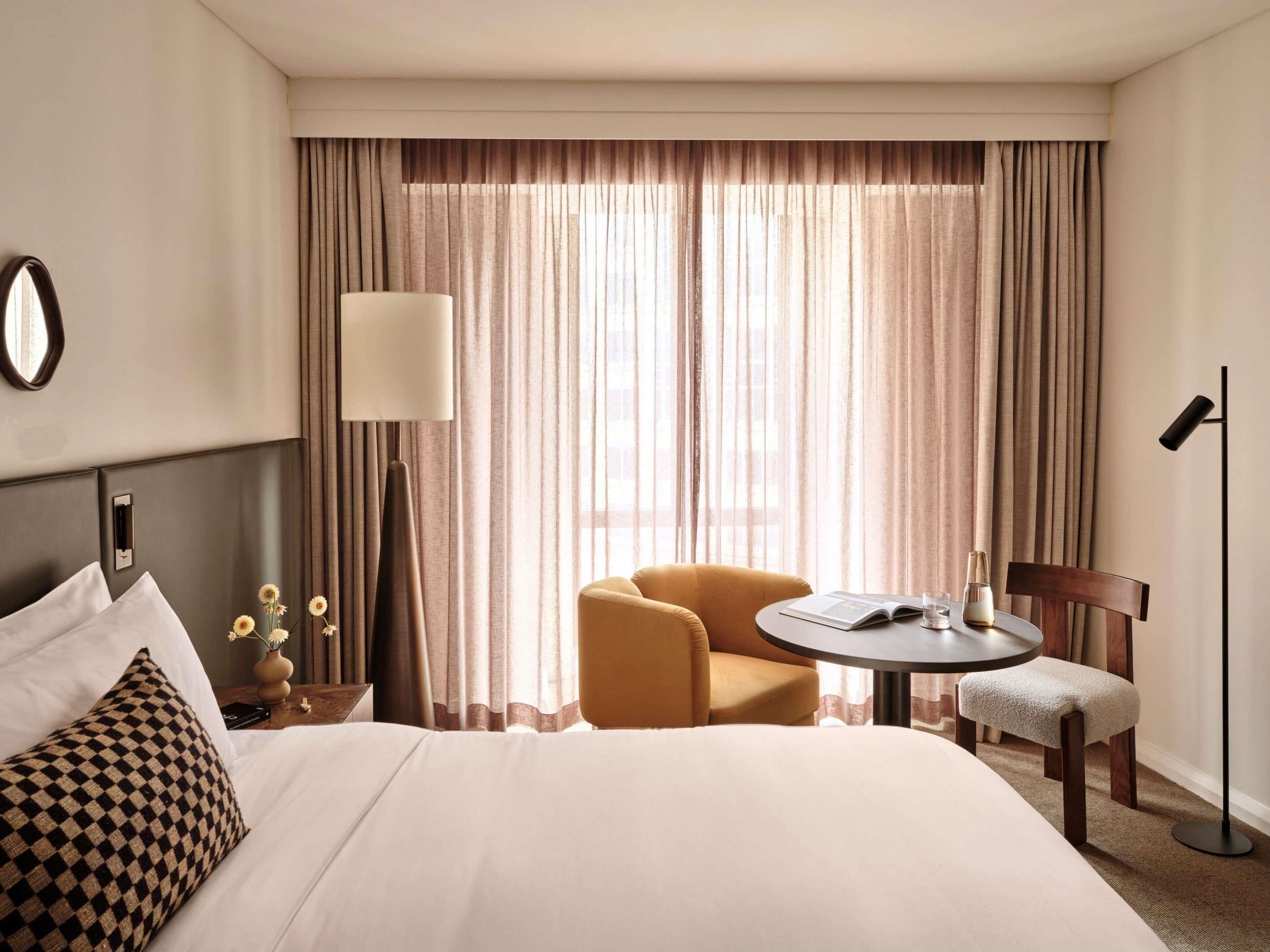 A hotel room with a bed, a round table, a chair, a lamp, and a large window with curtains.