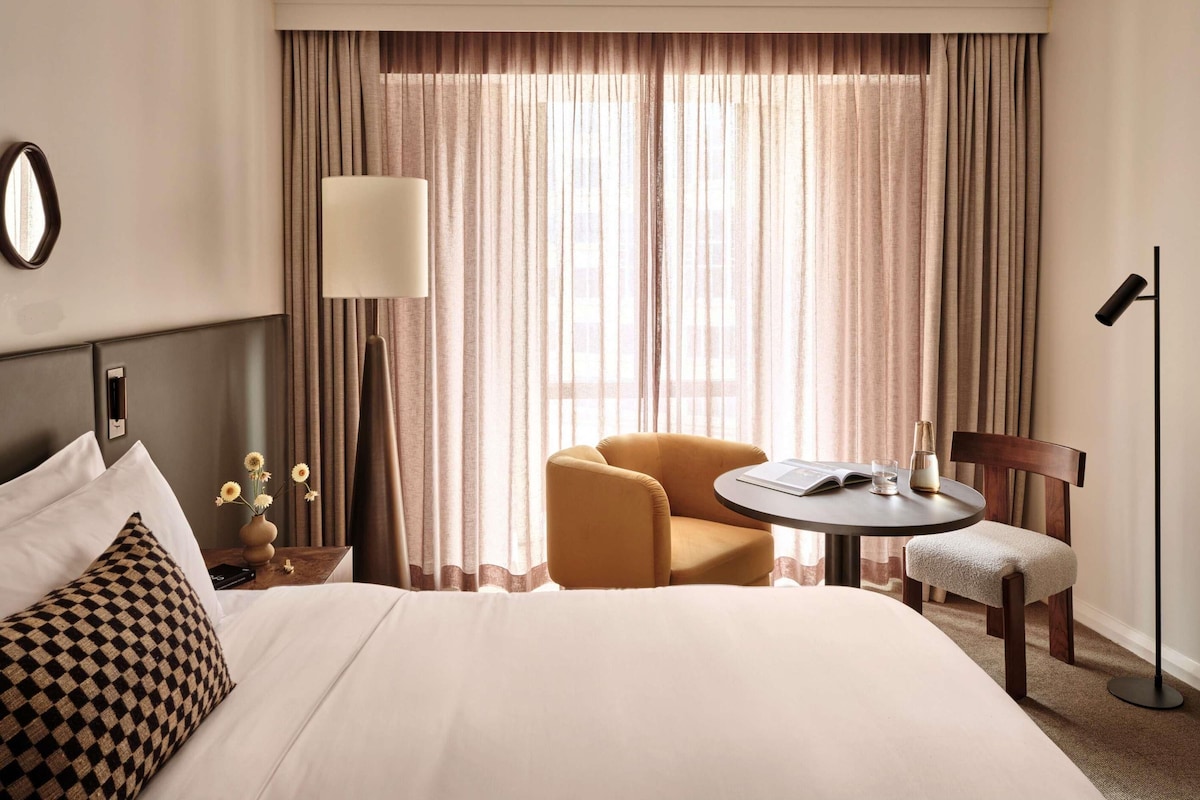 A hotel room with a bed, a round table, a chair, a lamp, and a large window with curtains.