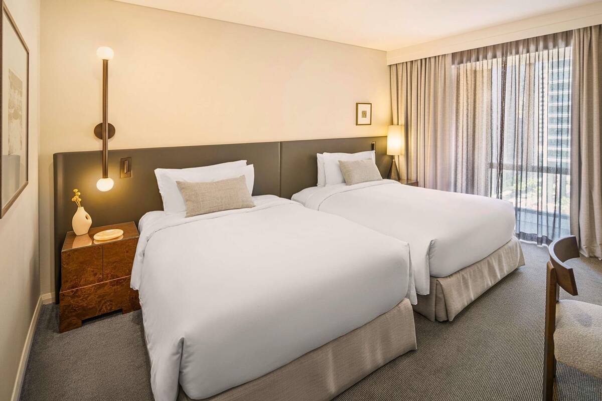 Ein Hotelzimmer mit zwei Betten, einem Nachttisch, einer Lampe, einem Stuhl und einem Fenster mit Vorhängen.