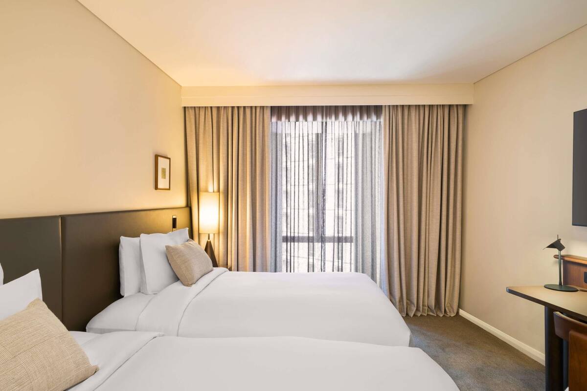 Ein Hotelzimmer mit zwei Betten, einem Schreibtisch, einer Lampe und einem großen Fenster mit Vorhängen.