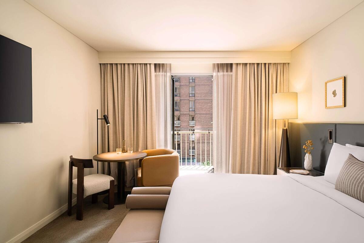 Ein Hotelzimmer mit Bett, Schreibtisch mit zwei Stühlen, Fernseher und einem Balkon mit Vorhängen.