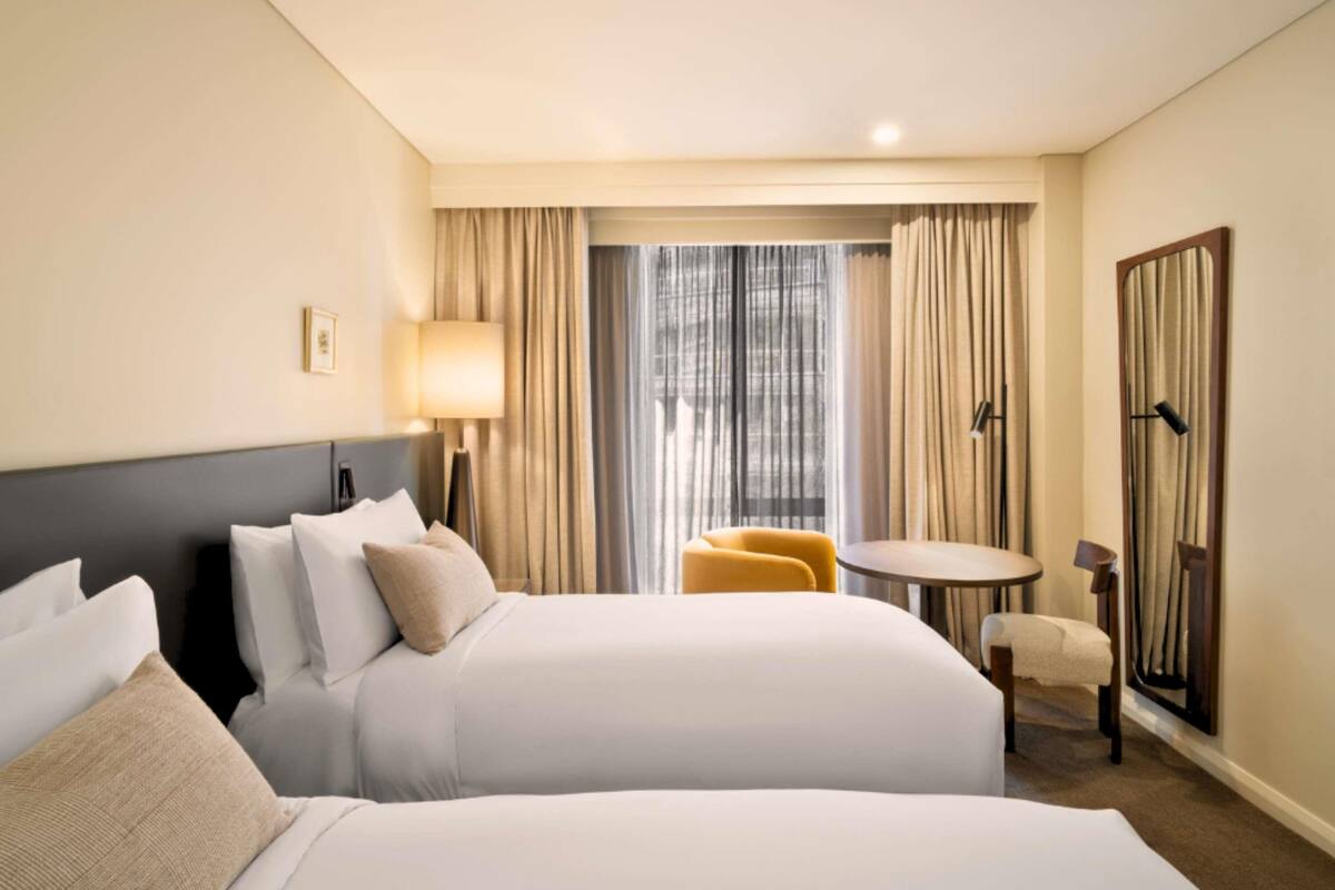 Ein Hotelzimmer mit zwei Betten, einem kleinen Tisch, einem Sessel und einer Lampe, mit Blick auf die Stadt durch das Fenster.