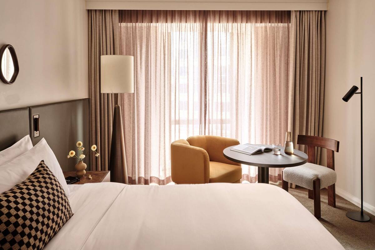 Ein Hotelzimmer mit einem Bett, einem runden Tisch, einem Stuhl, einer Lampe und einem großen Fenster mit Vorhängen.