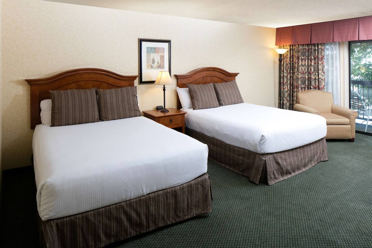 A hotel room with two beds, a nightstand with a lamp, a chair, and a window with curtains.