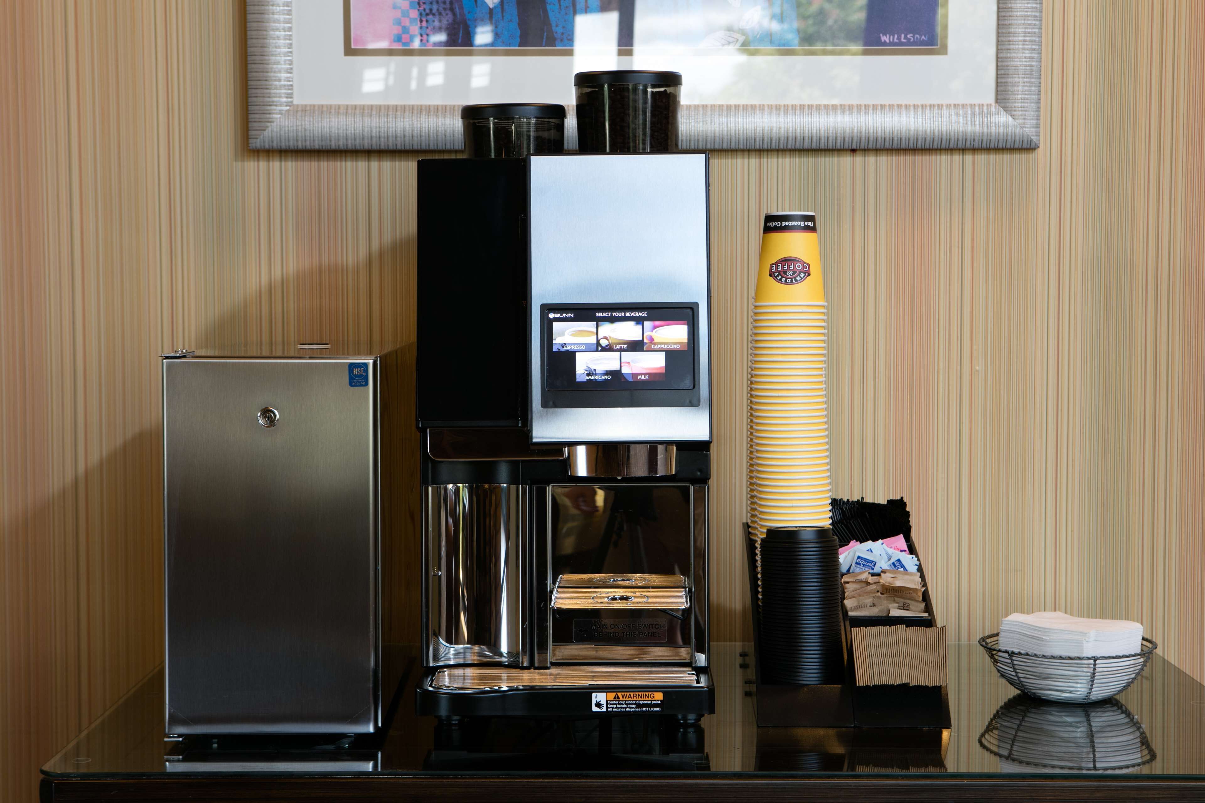 Una cafetera con pantalla digital, un refrigerador pequeño y una pila de vasos desechables.
