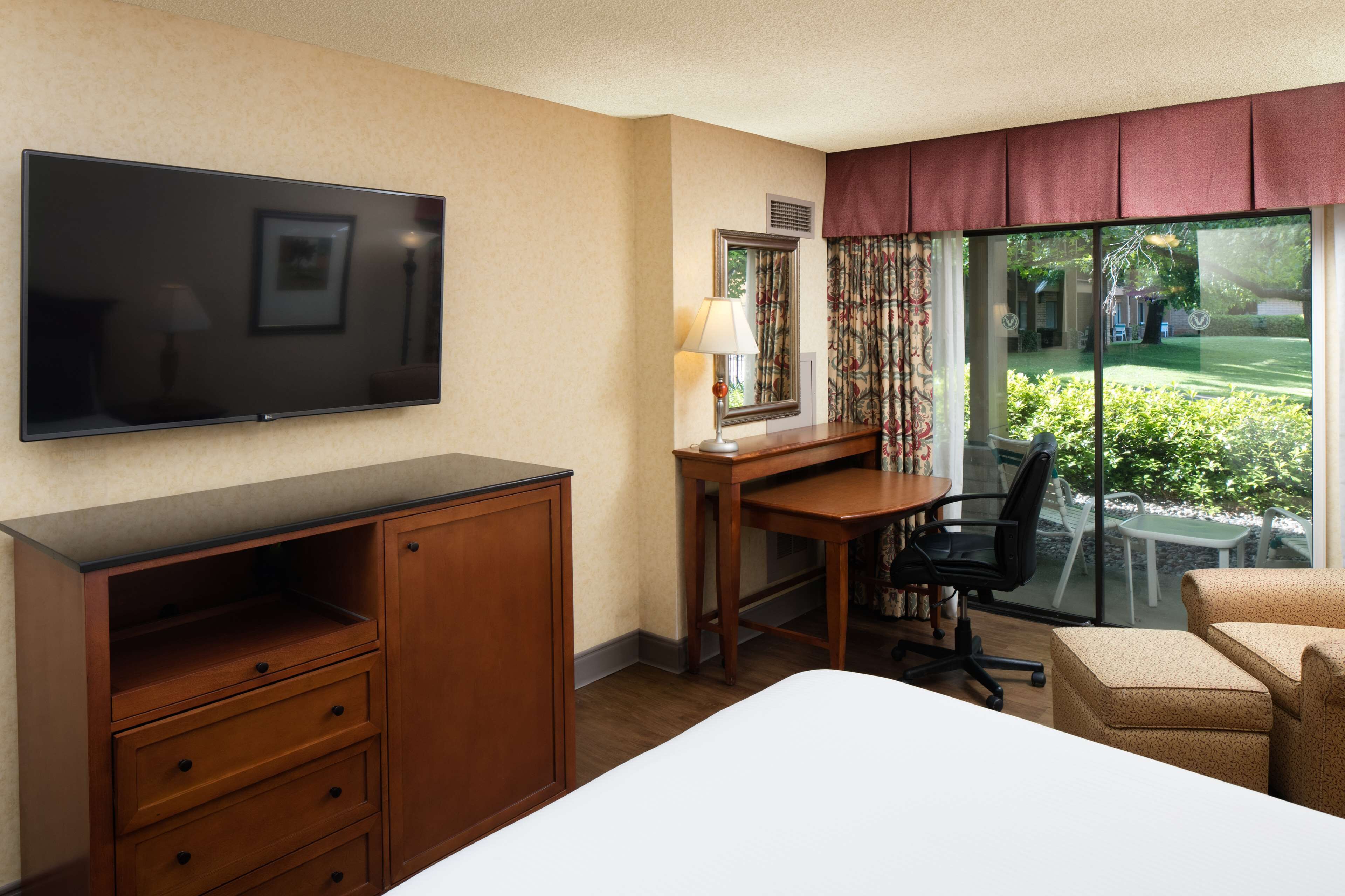 A hotel room with a bed, a desk with a chair, a television, and a window with a view of greenery.