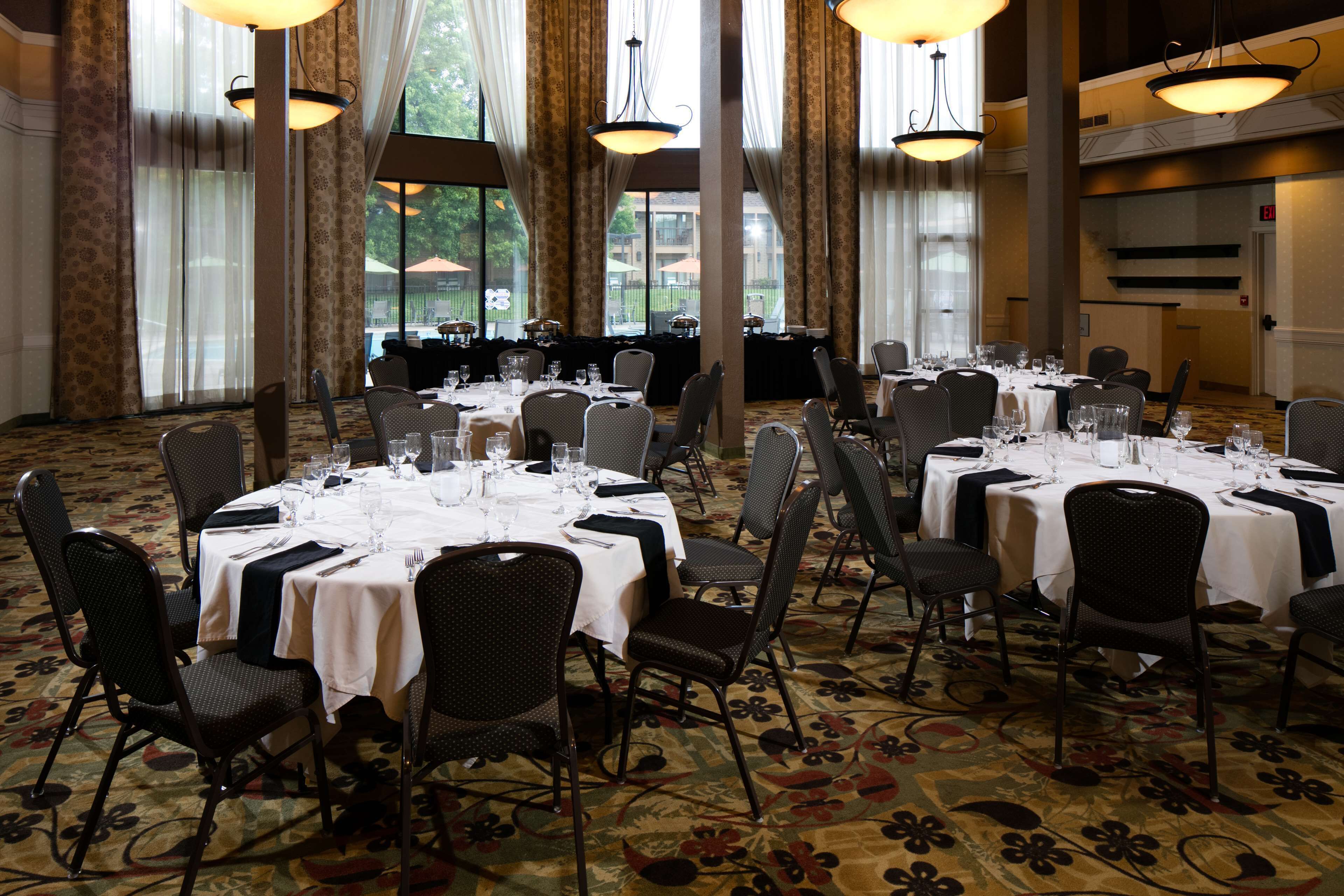 A banquet hall with multiple round tables set for dining, large windows, and a carpeted floor.