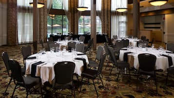 A banquet hall with multiple round tables set for dining, large windows, and a carpeted floor.