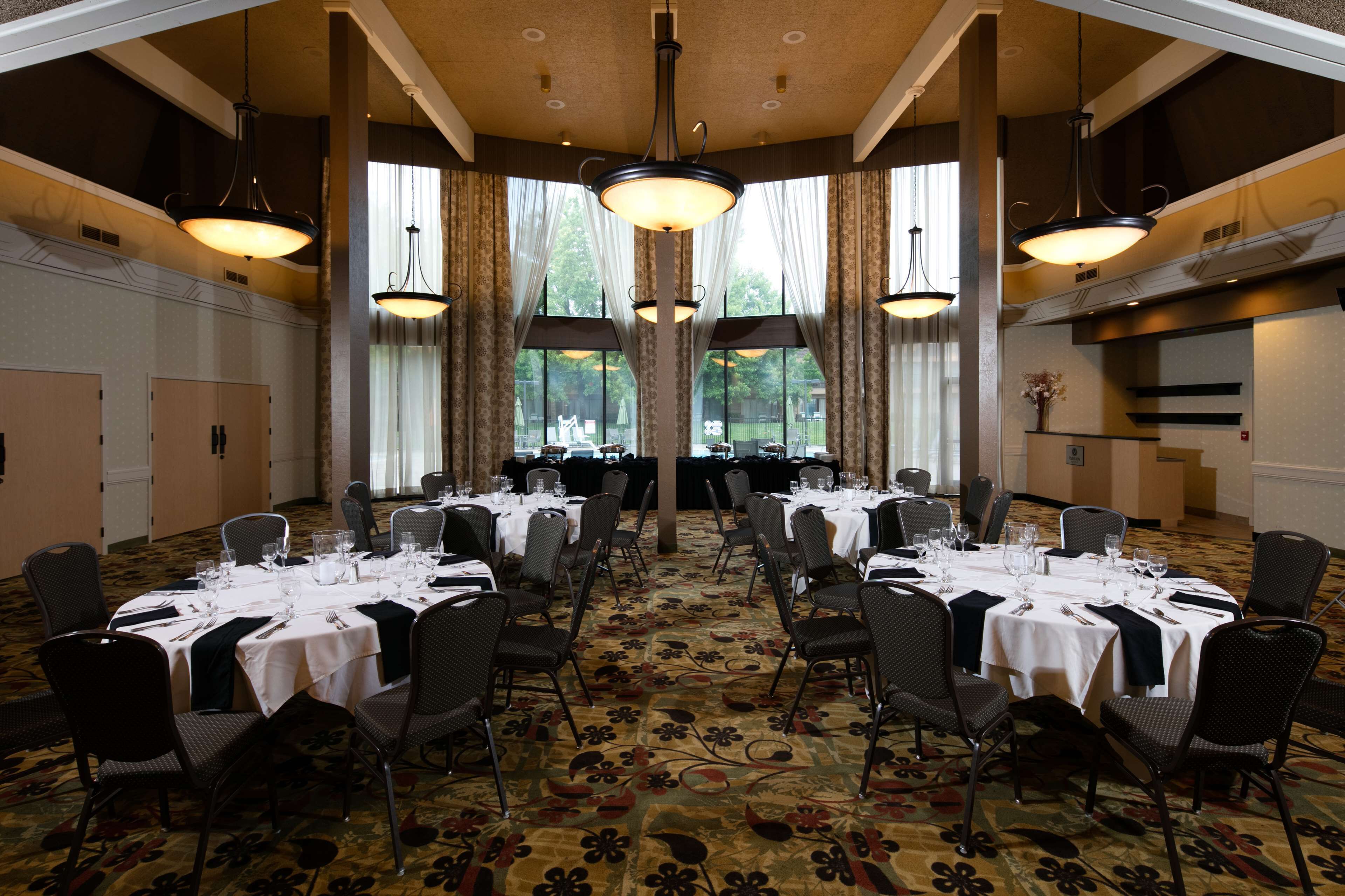 Une salle de banquet avec des tables rondes dressées pour le repas, de grandes fenêtres et des lustres.