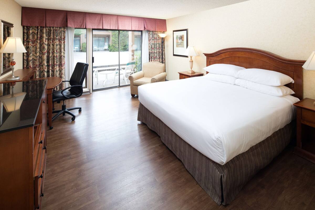 A hotel room with a large bed, a desk, a chair, and a view of the outdoors through a sliding door.