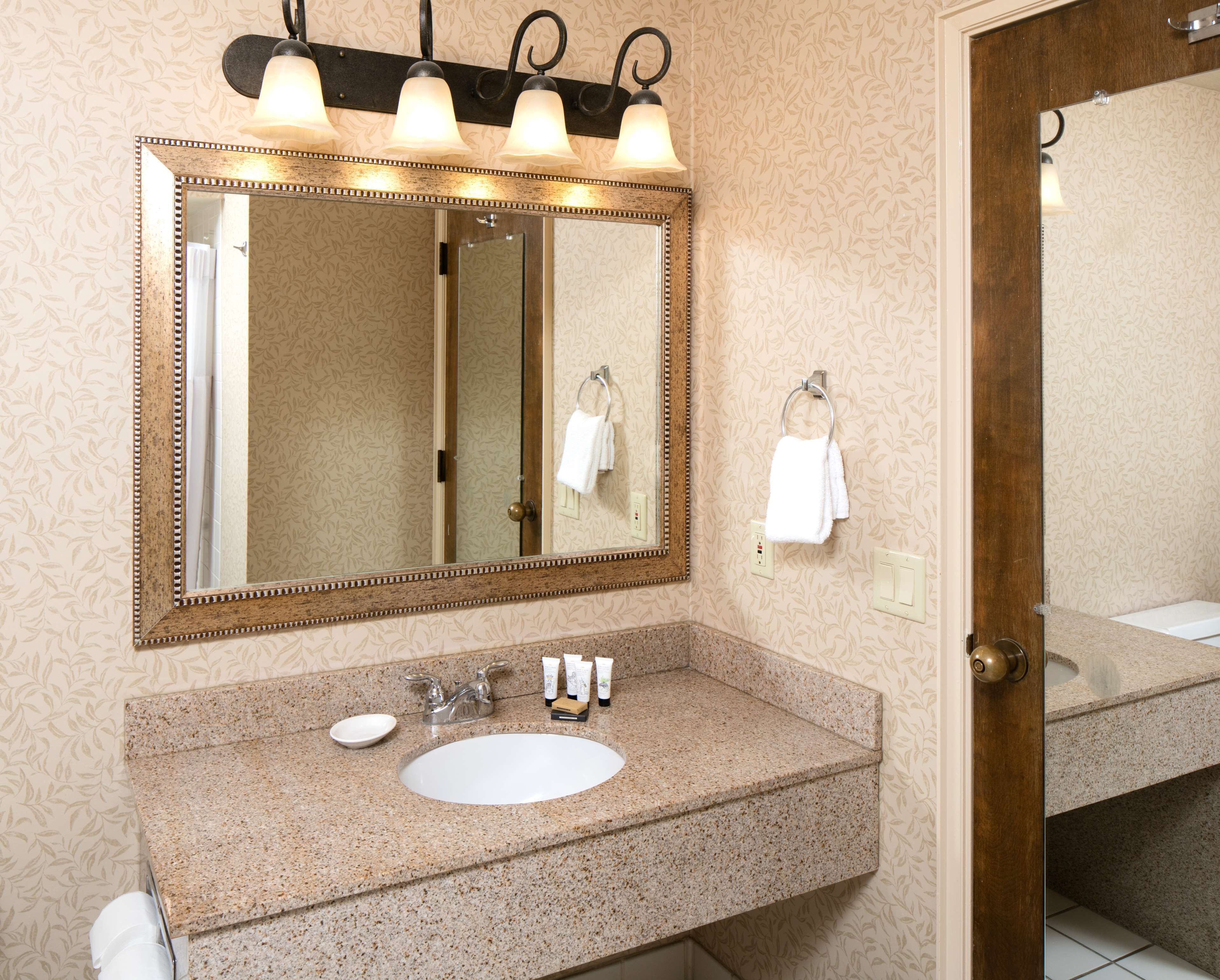A bathroom with a granite countertop sink, a large mirror with three lights, and two towels hanging on hooks.
