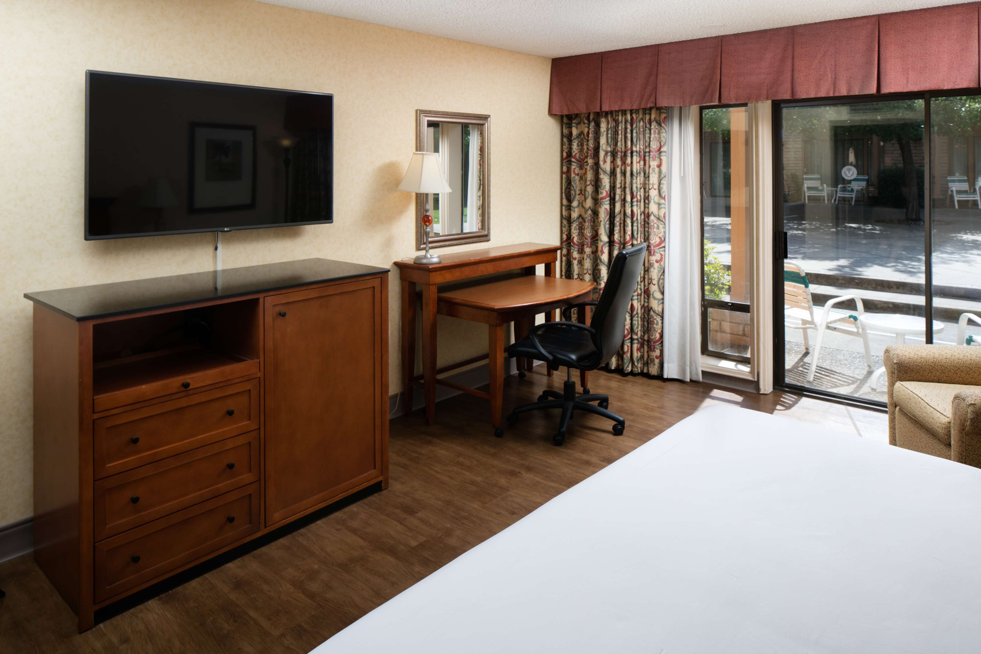 A hotel room with a bed, a desk, a chair, a TV, a dresser, and a sliding glass door to the outside.