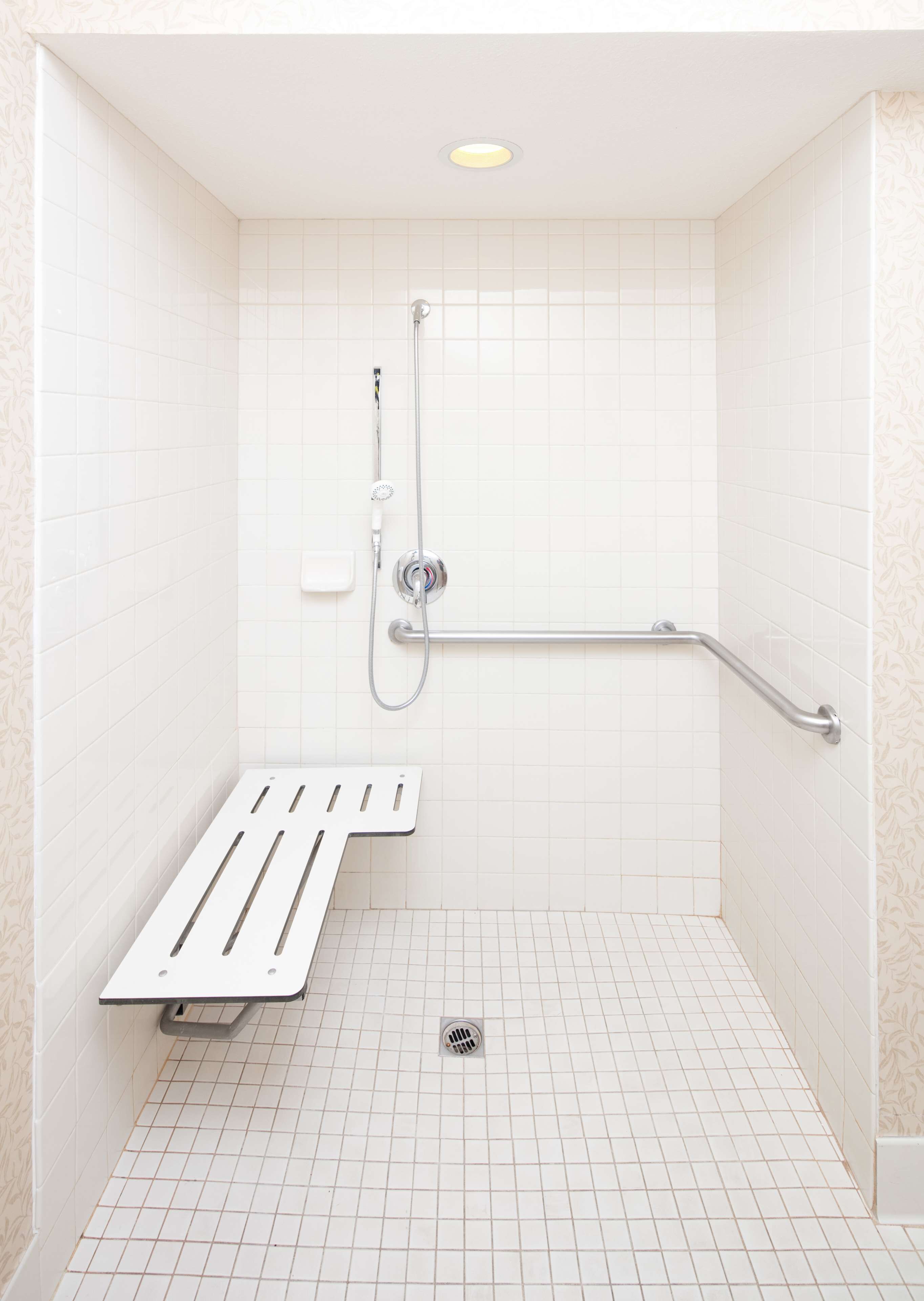 A small, white-tiled shower room with a grab bar and a bench.